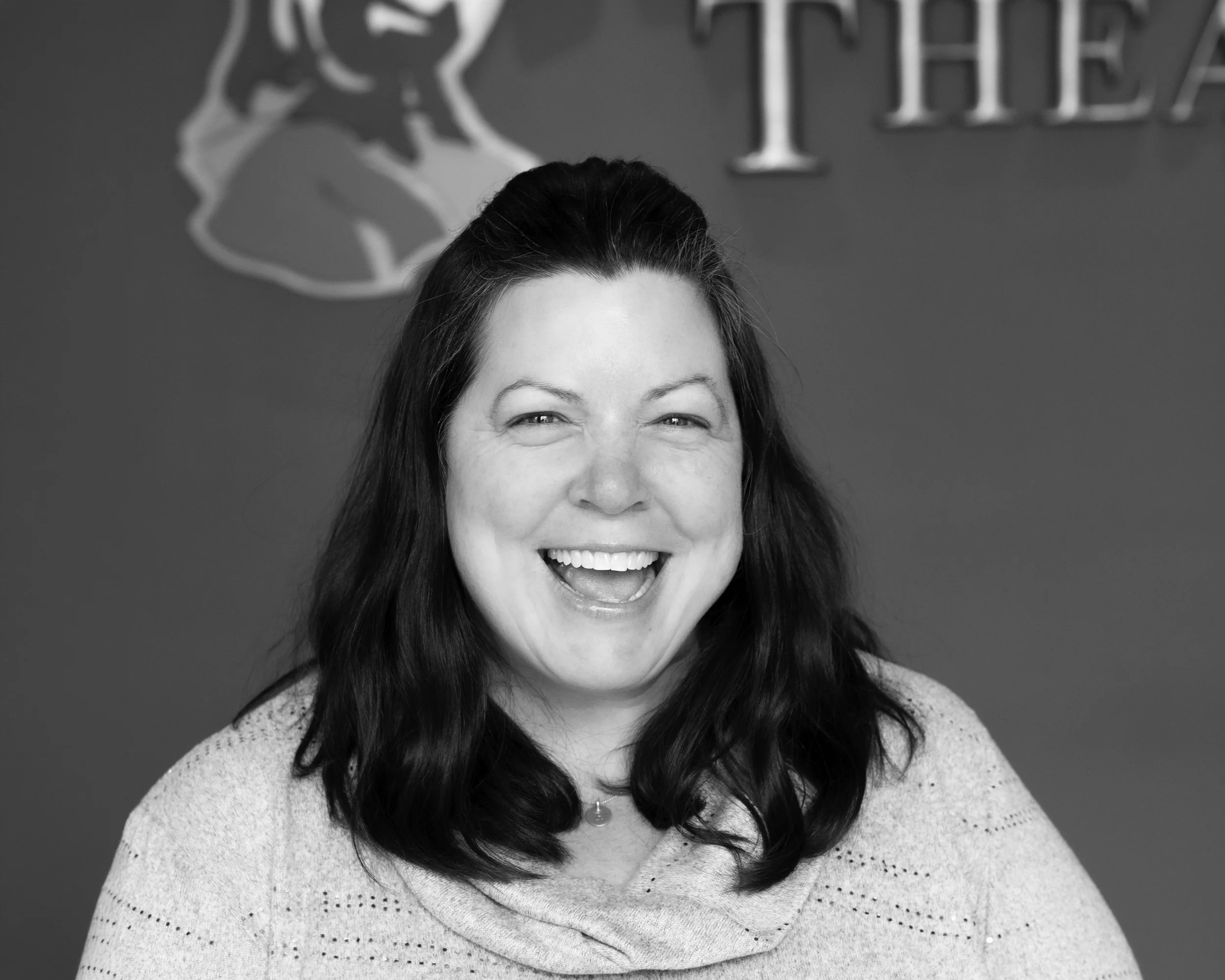 A black-and-white photo of a smiling woman with shoulder-length dark hair, wearing a light-colored sweater, in front of a wall with the partial word 'HEALTH' and a lion illustration.