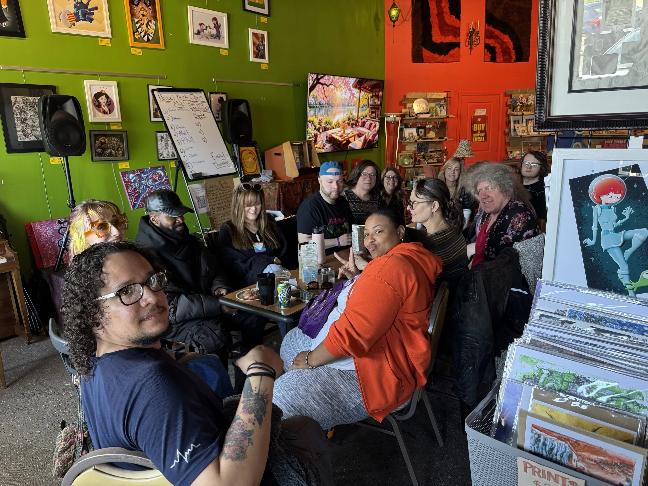 Group of people sitting at tables in an art gallery or cafe with colorful walls, framed artwork, and a whiteboard with notes in the background.