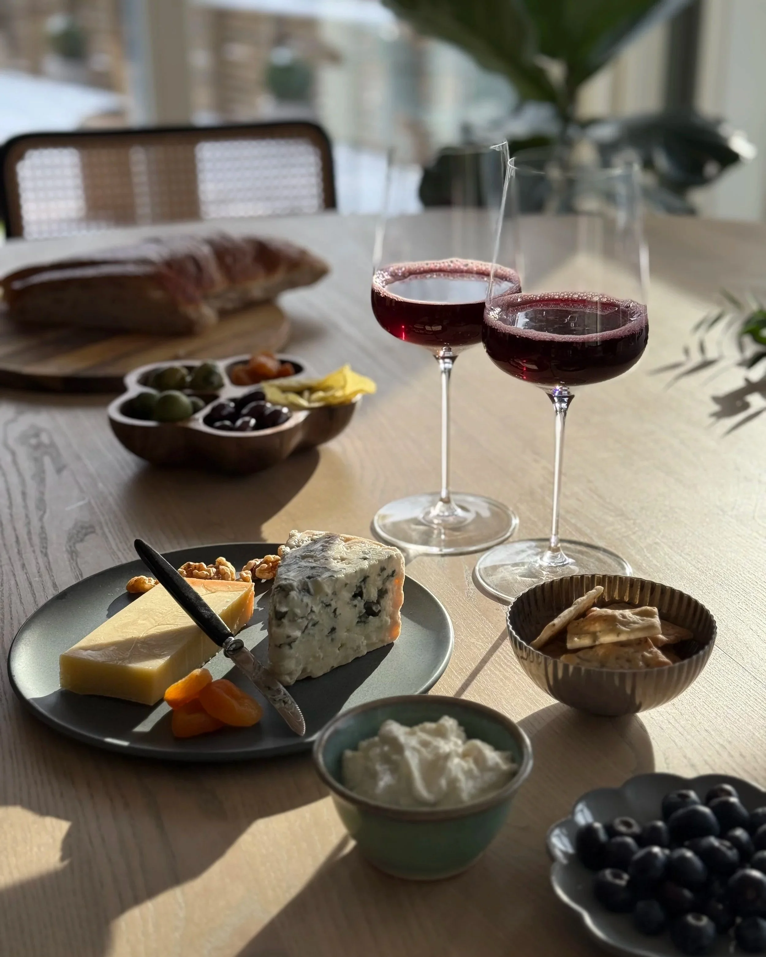 A table set with a variety of cheeses, fruits, and beverages, including two glasses of red wine. The background shows a window and some indoor plants.