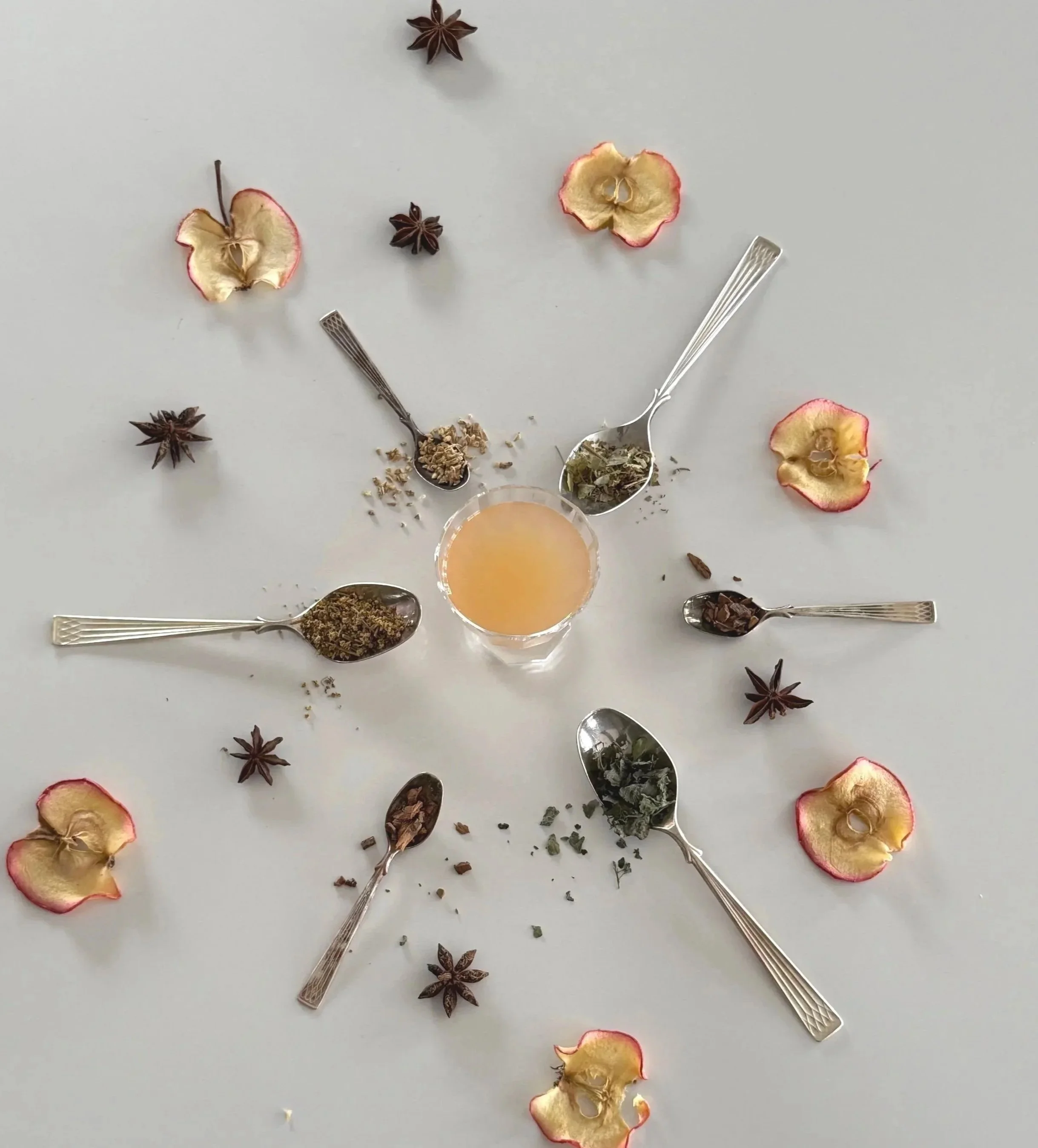 Flat lay photo of dried apple slices, star anise, dried herbs, and spoons containing dried spices and herbs on a white surface, with a glass of Ravn & By sparkling botanical drink No 1, light coloured, drink in the center.