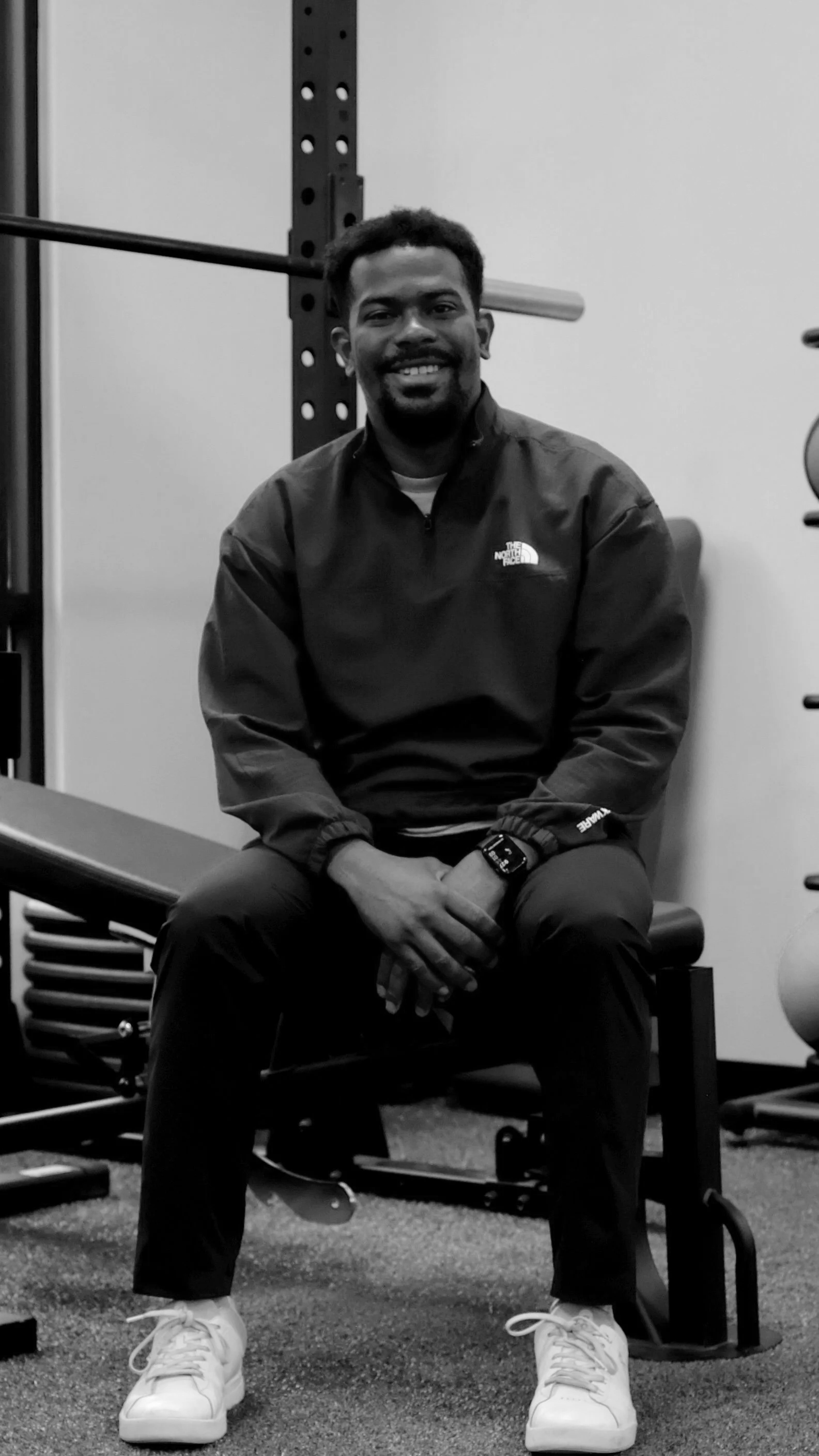 A smiling man sitting on a bench in a gym, wearing a black North Face jacket, dark pants, white sneakers, and a watch, with gym equipment in the background.