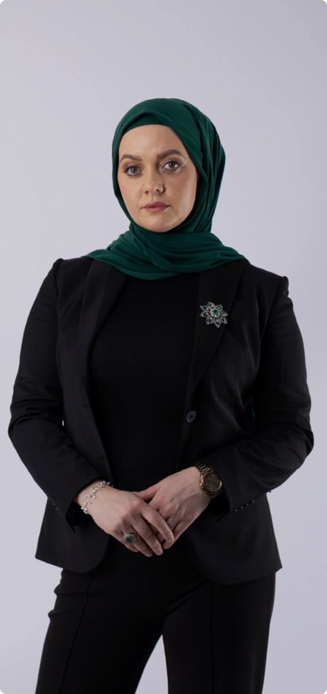 A woman wearing a dark blazer, black top, green headscarf, and jewelry poses against a plain gray background.