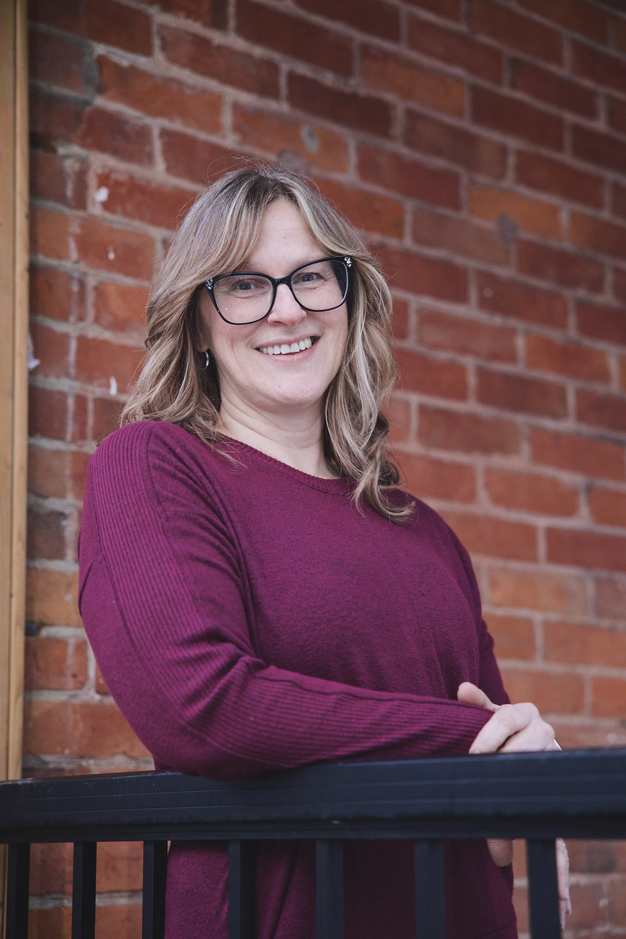 A woman with blonde hair, wearing glasses and a maroon sweater, smiling and standing in front of a brick wall.