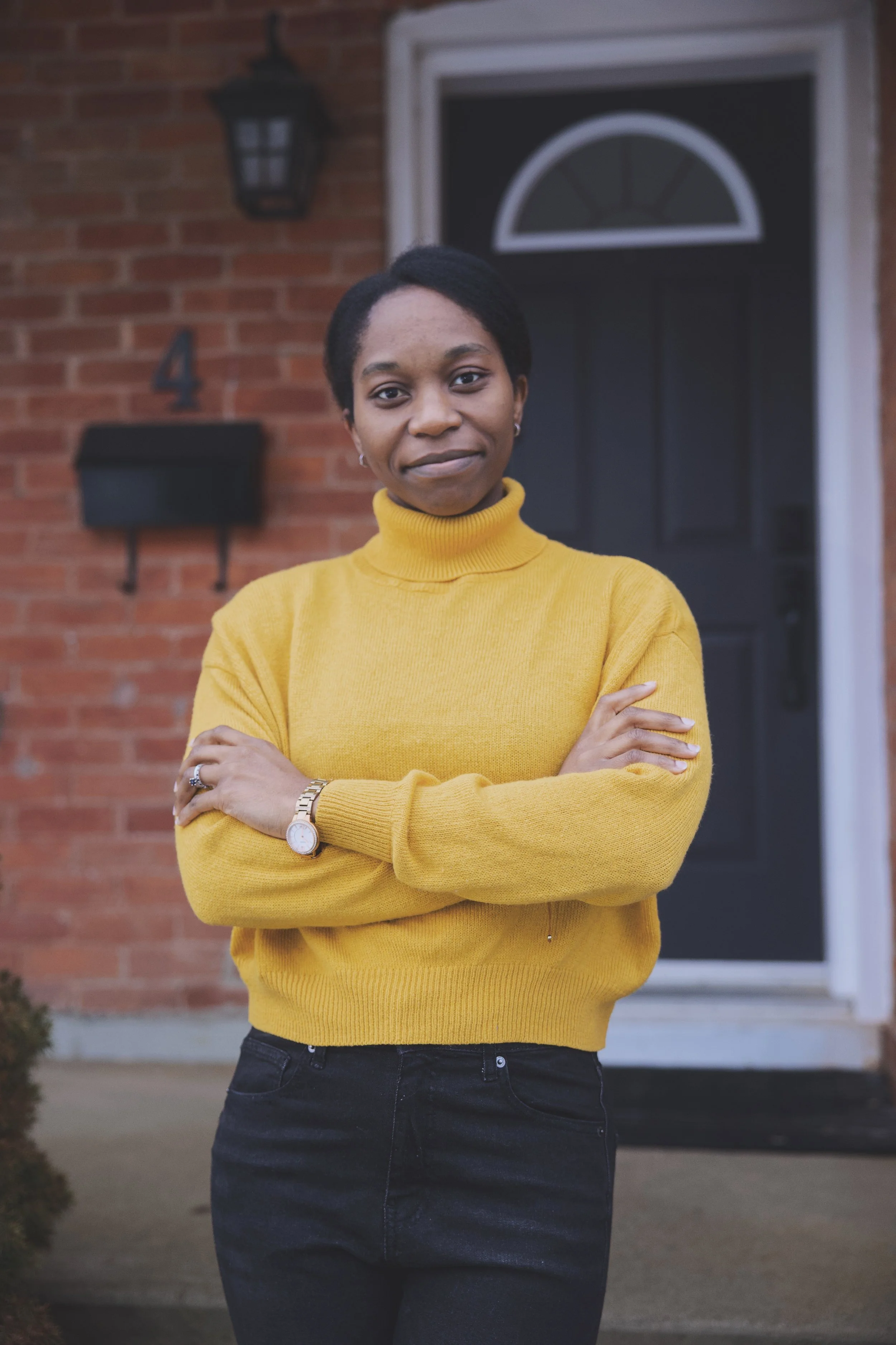 A woman standing in front of a house with her arms crossed, wearing a yellow sweater and black pants.