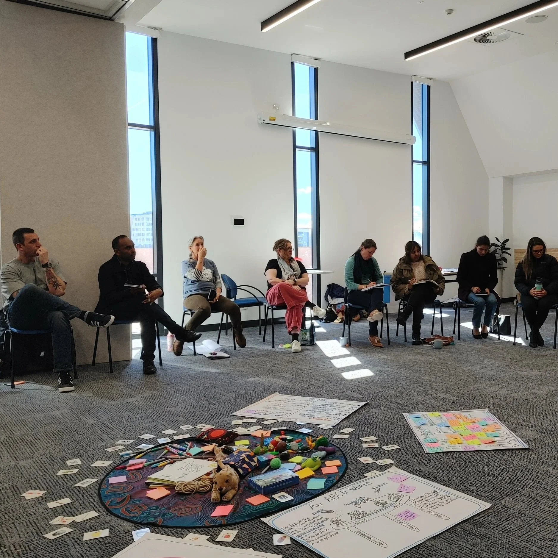 People seated in a semi-circle in a bright conference room, with materials and toys on the floor, participating in a group activity or workshop.