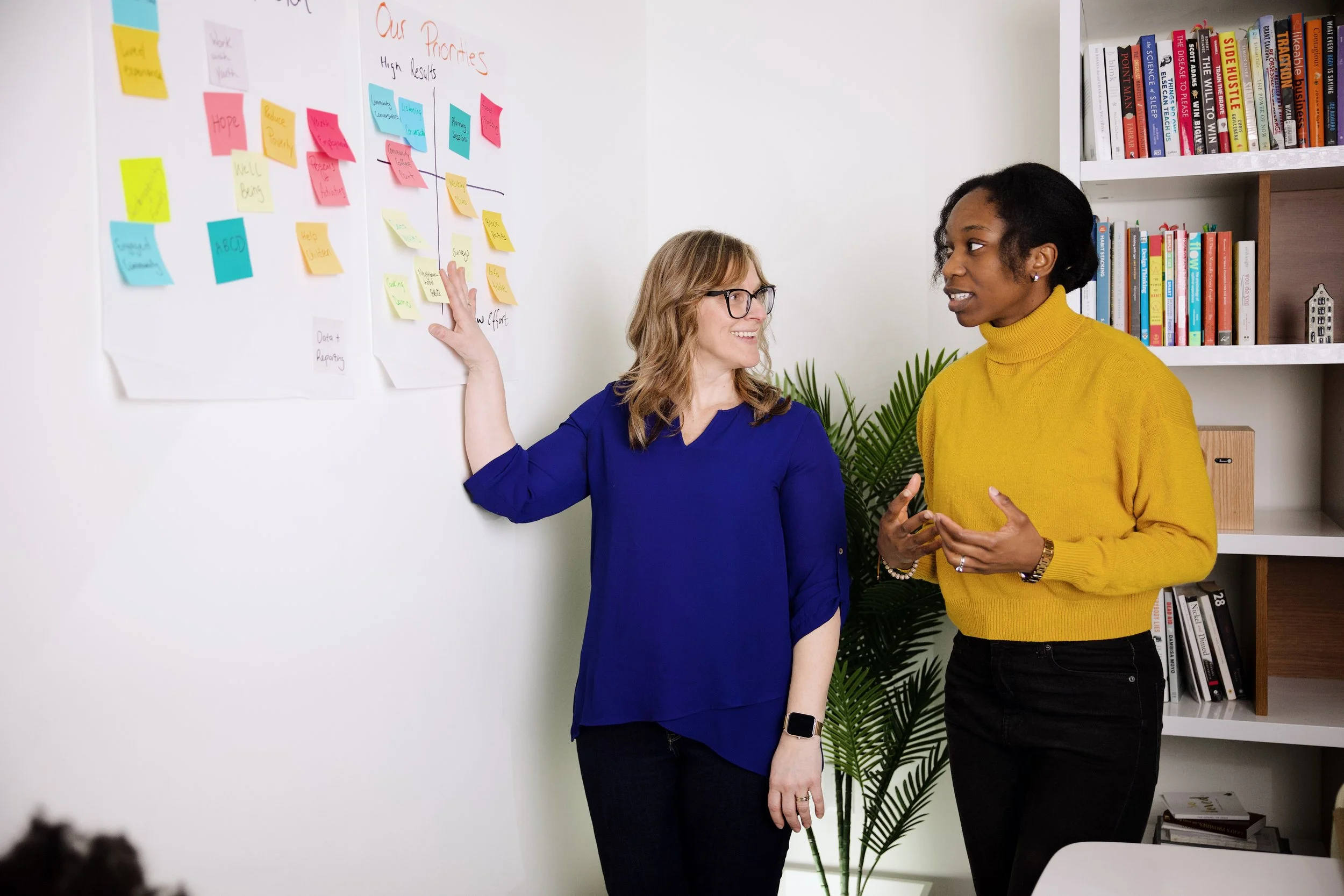 Two women standing and talking in an office, one points at a whiteboard with colorful sticky notes, and a bookshelf in the background.