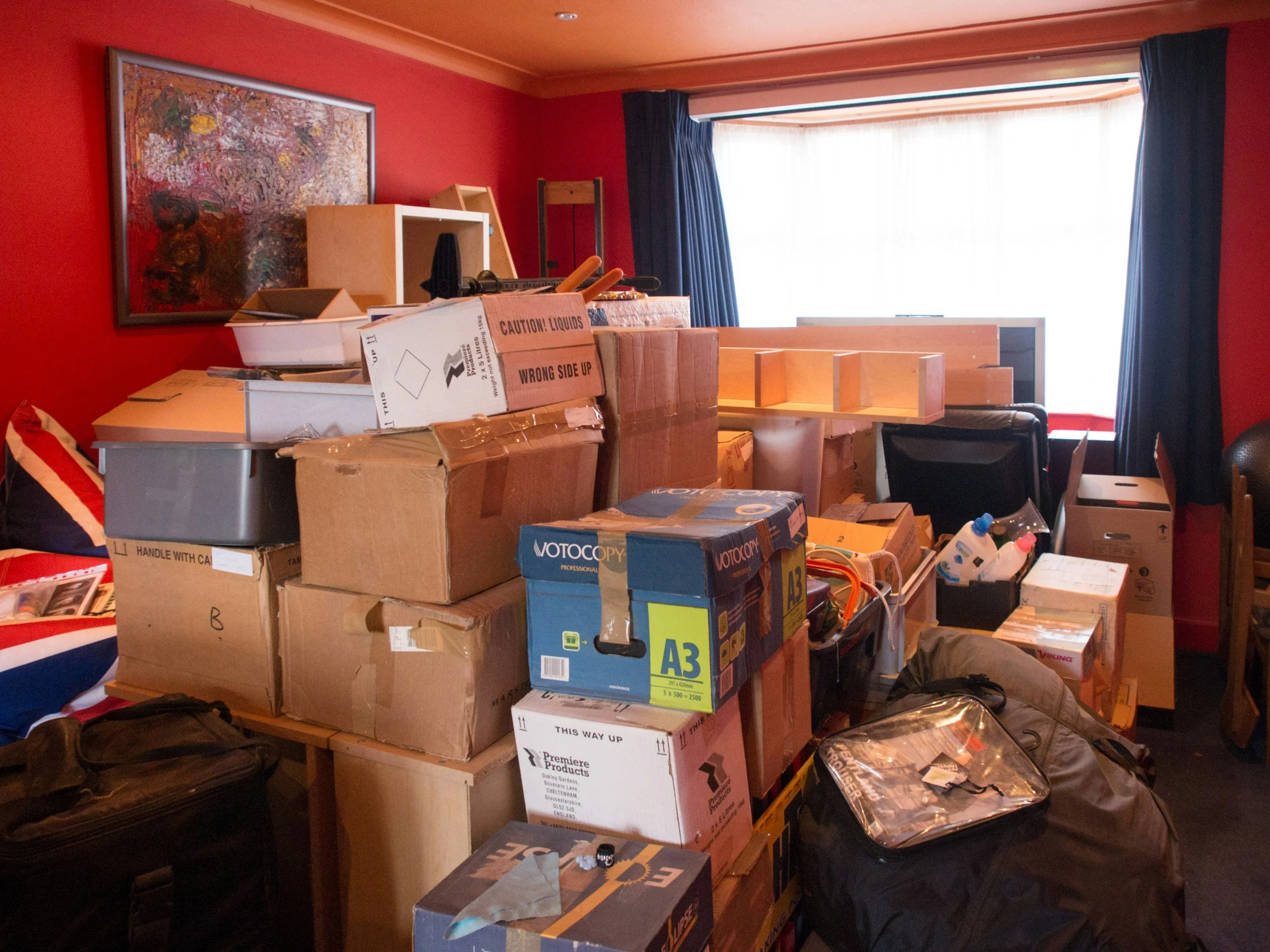Room filled with scattered boxes, some labeled and others plain, and various items including a black bag, a chair, and a bag of bottles near a window with dark curtains, red walls, and artwork on the wall.