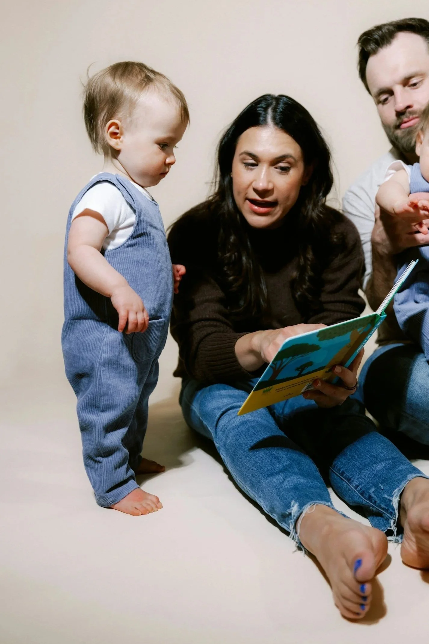 A woman sitting on the floor reading a children's book with a little boy standing beside her. In the background, a man is holding a young child, all looking at the book.