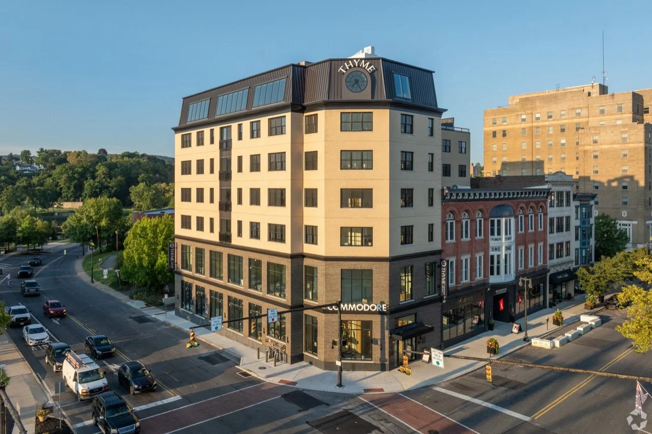 A multi-story building on a street corner with storefronts at the ground level, surrounded by trees and cars, under a clear sky. The Commodore Easton
