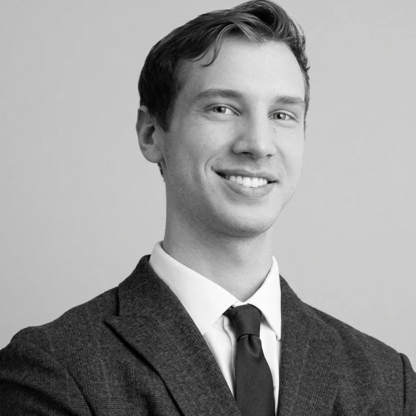 Black and white portrait of a young man with short dark hair, smiling, wearing a suit with a white shirt and a dark tie, against a plain background.