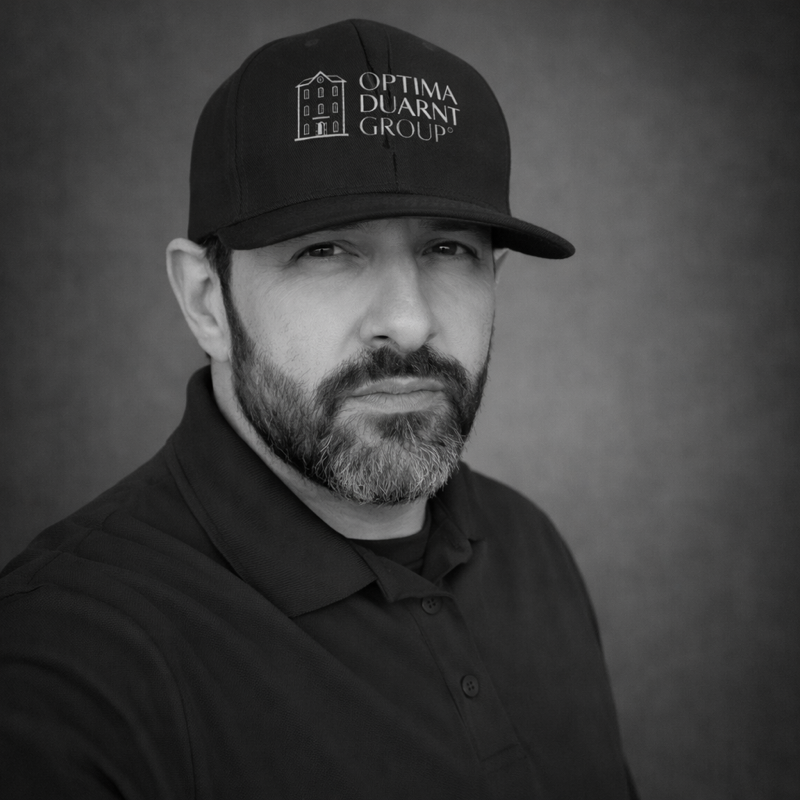 Black and white portrait of a man with a beard wearing a cap with the logo and text 'Optima Duarnt Group' and a collared shirt.