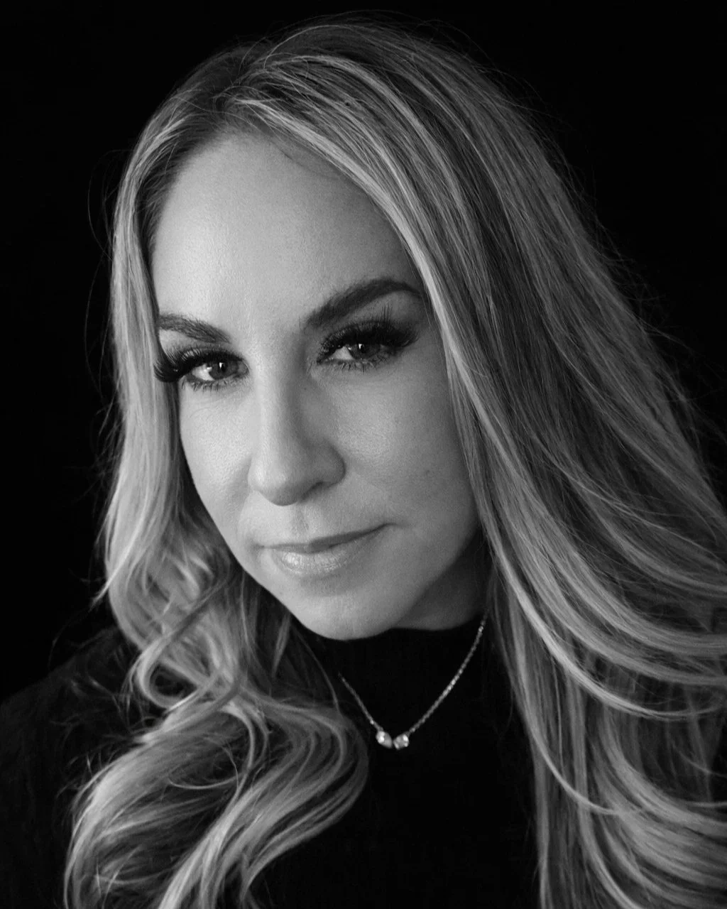 Black and white portrait of a woman with long, wavy hair, wearing makeup and a necklace, looking at the camera.