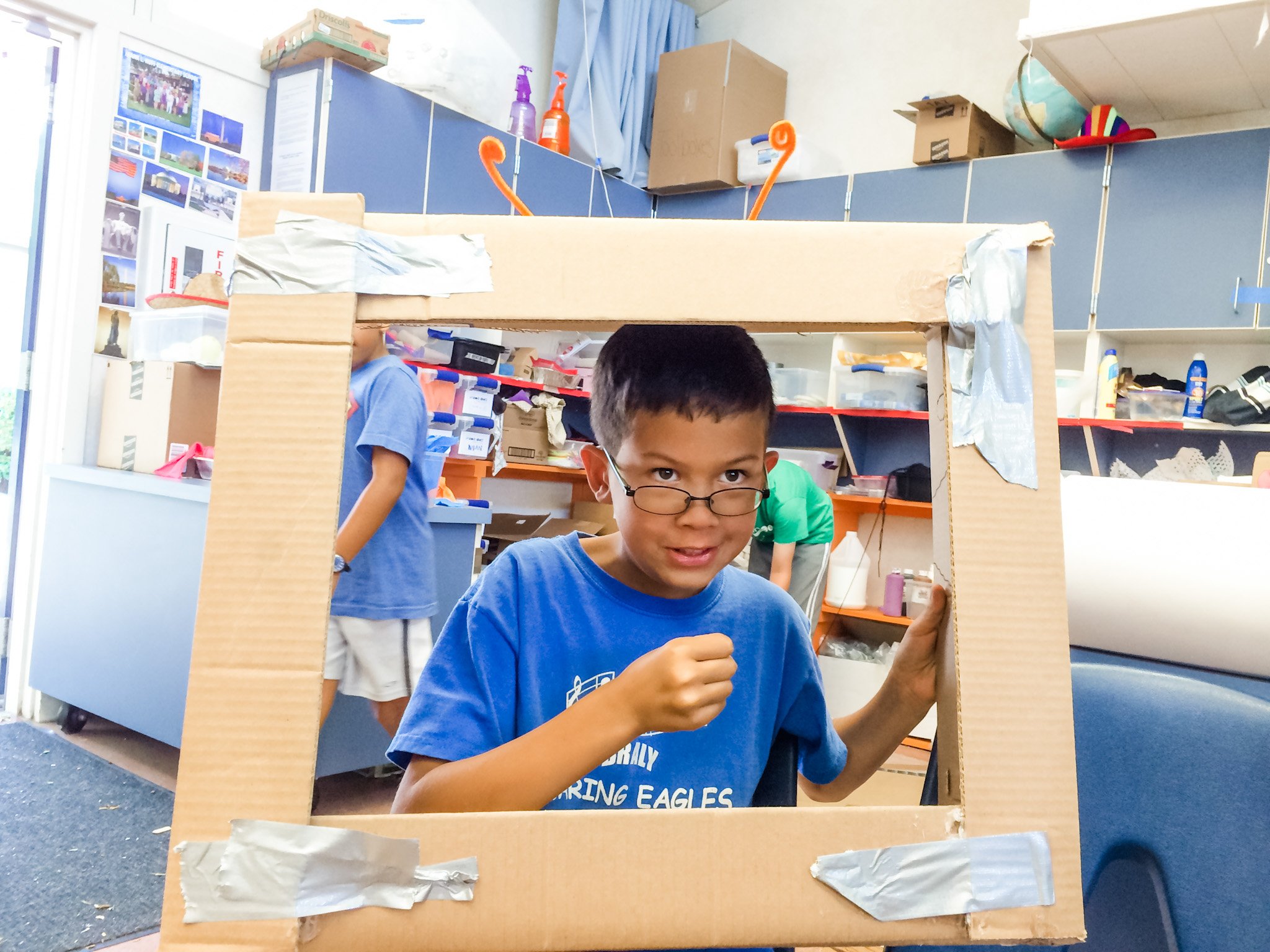 A young boy with glasses and a blue t-shirt is holding a large cardboard frame and looking through it, with a classroom setting in the background.