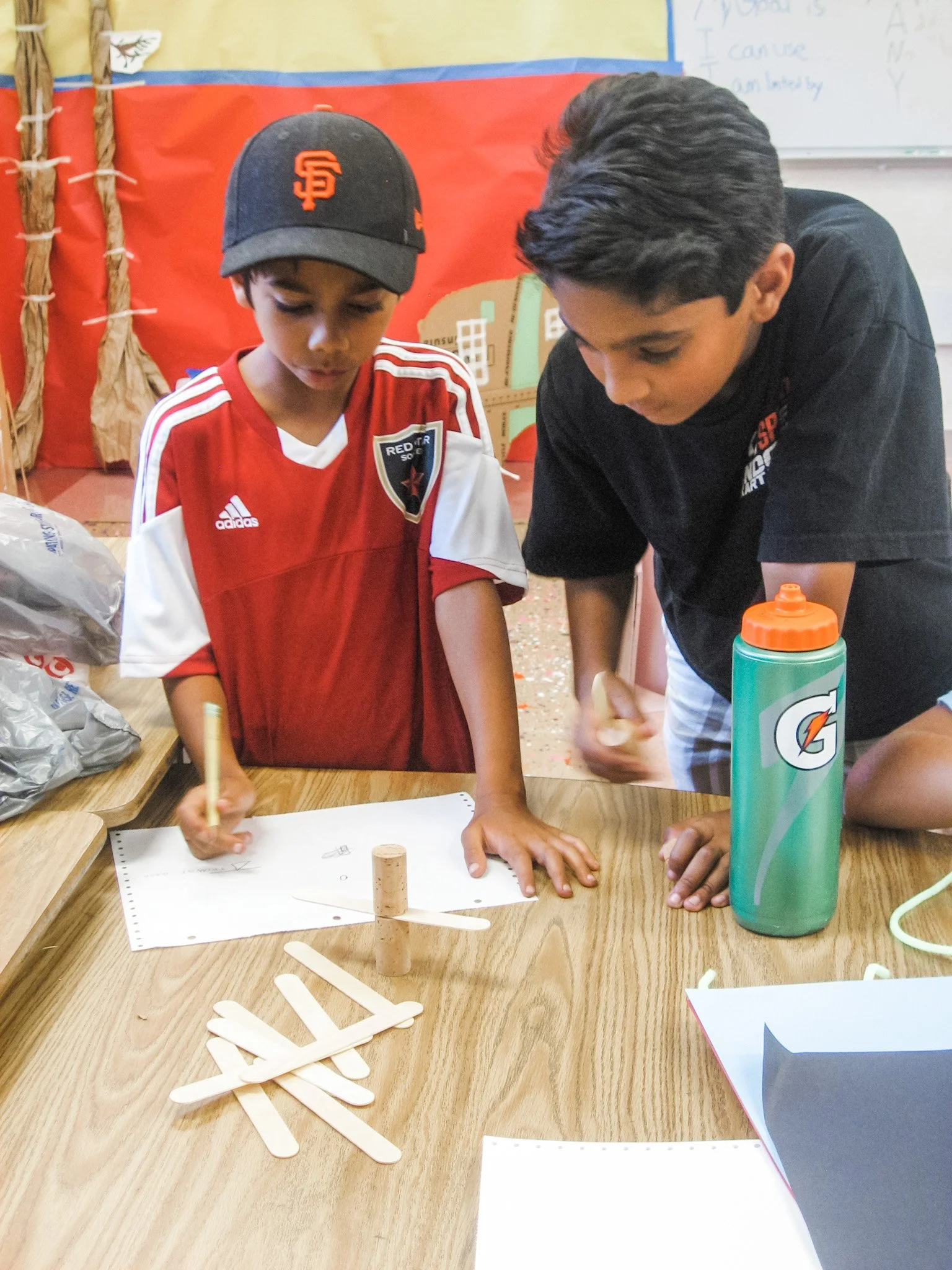 Two boys working together on a school project at a wooden table, with craft sticks and a cork, in a classroom setting.