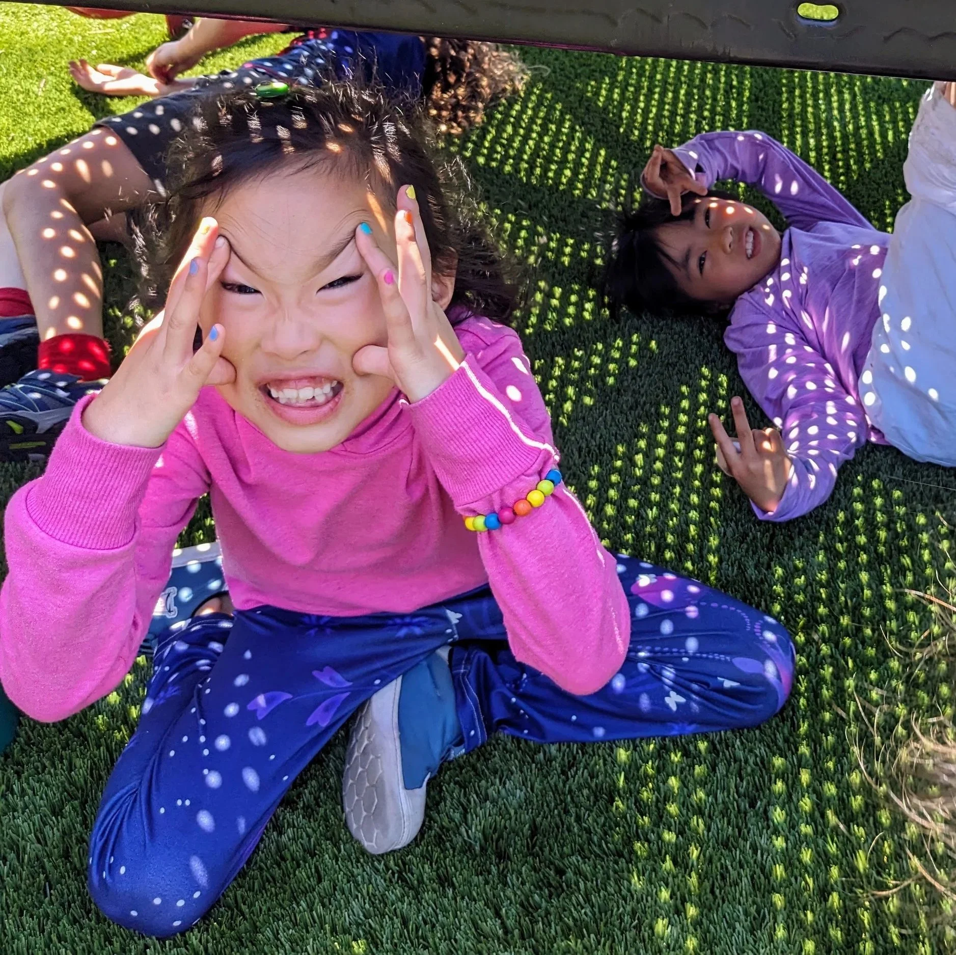 Two young girls lying on the ground under a shade structure, one making a funny face and the other smiling, both with playful poses.