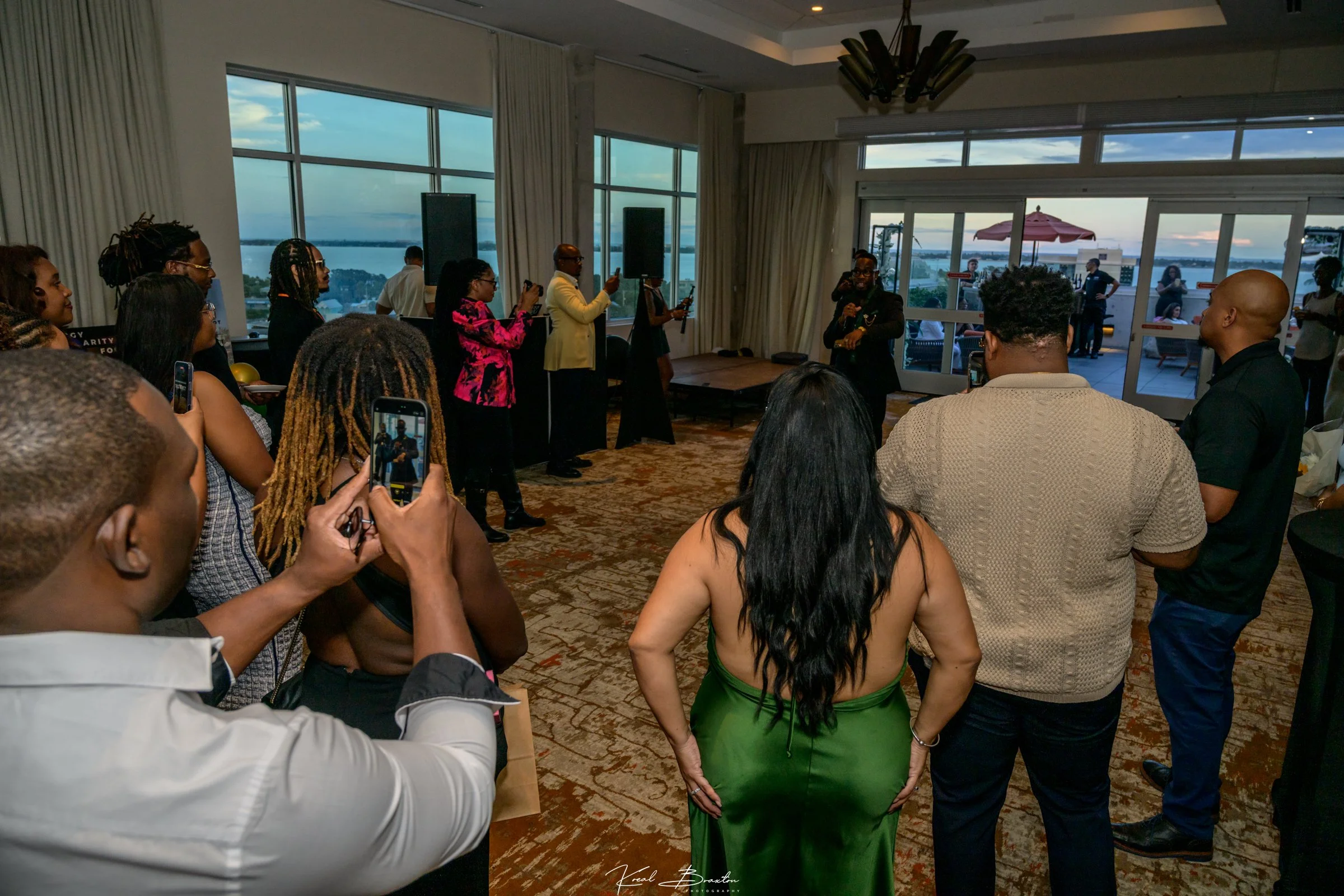 A group of people gathered indoors, some taking photos or recording a woman who is speaking into a microphone. Large windows behind them show an outdoor patio with an umbrella and a view of the city skyline and water.