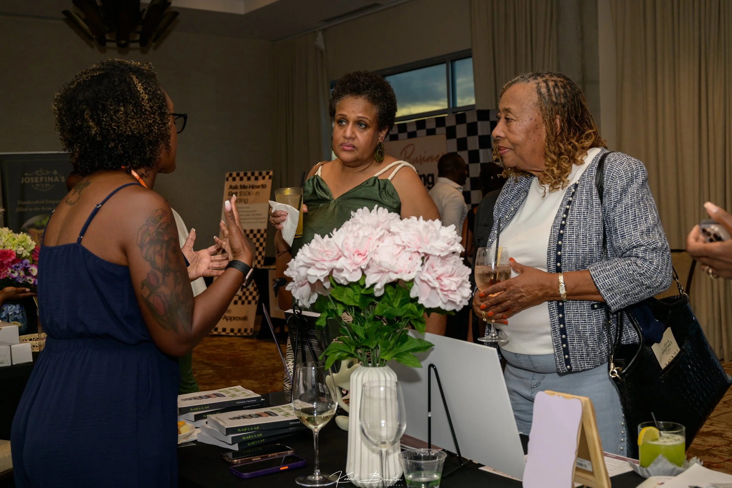 Three women engaged in conversation at a table. The table has a white flower arrangement, wine glasses with beverages, and stacked books. In the background, there are promotional posters and a window with curtains.
