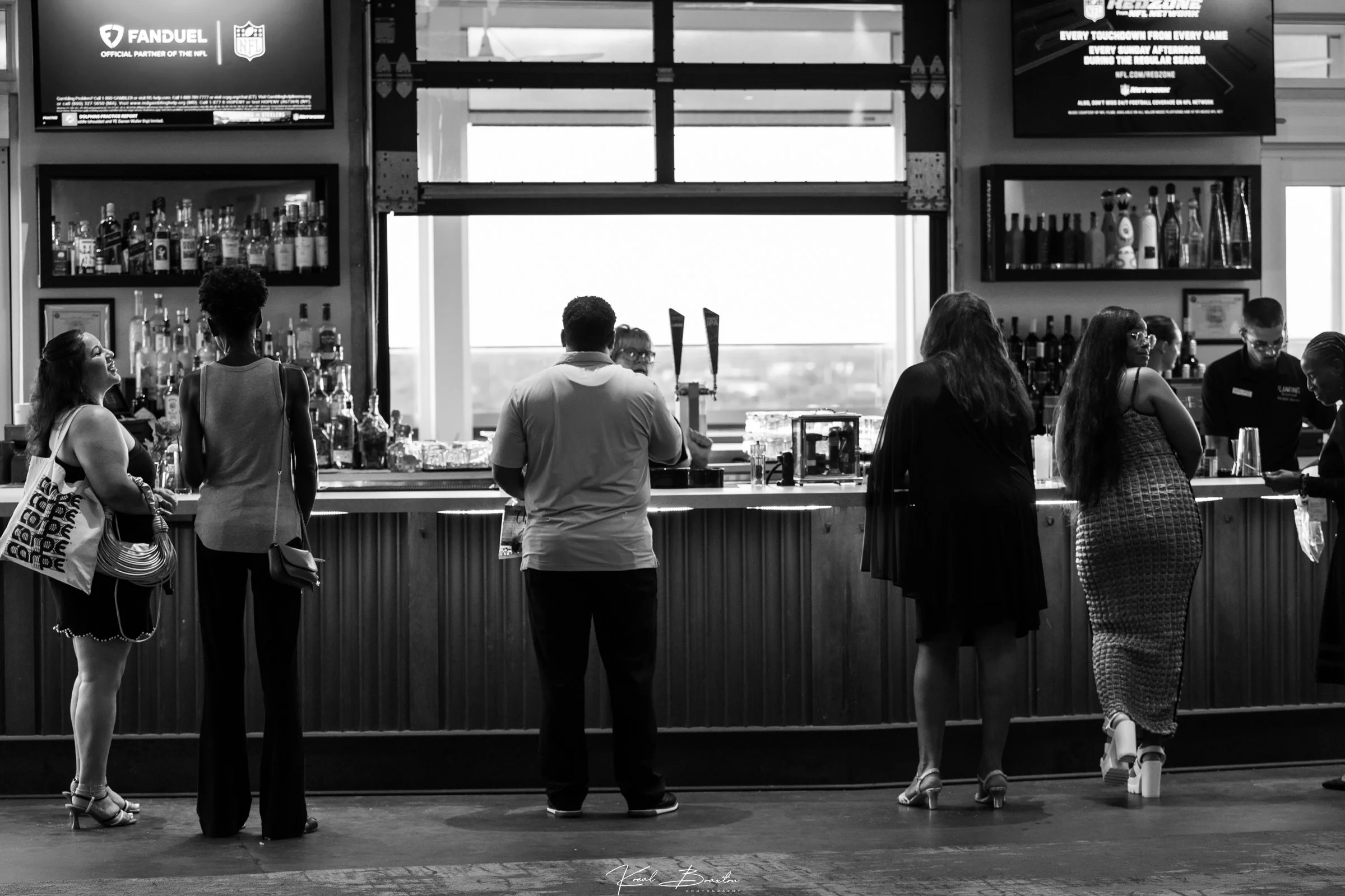People waiting in line at a bar or restaurant counter, with bottles and drinks visible behind the counter, and large screens displaying advertisements or information above. The scene is in black and white.