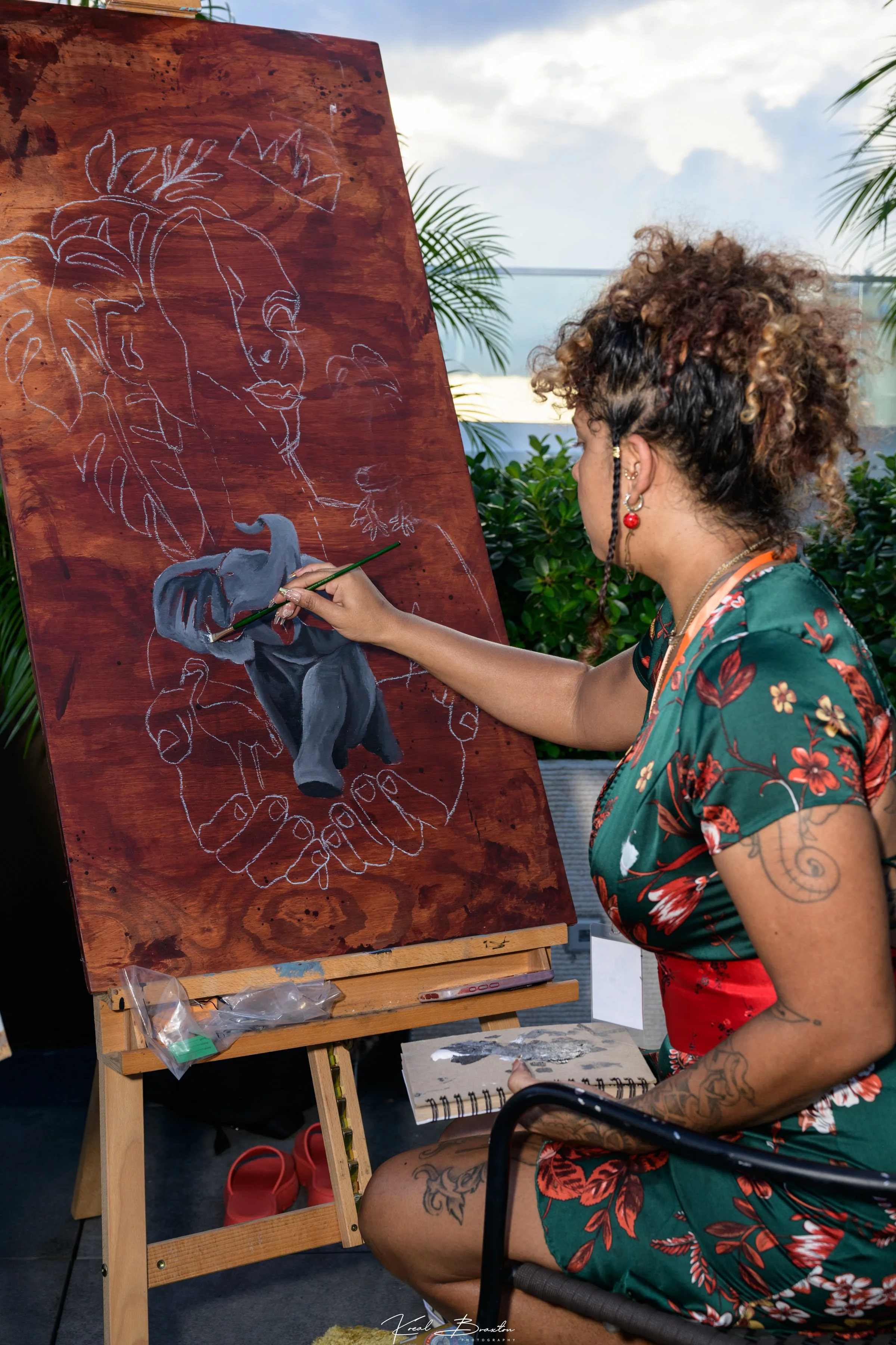 A woman with curly hair and tattoos is sitting in a chair while painting a portrait of a woman with large leaves in her hair on a large wooden canvas. She is wearing a floral dress and earrings with red beads.