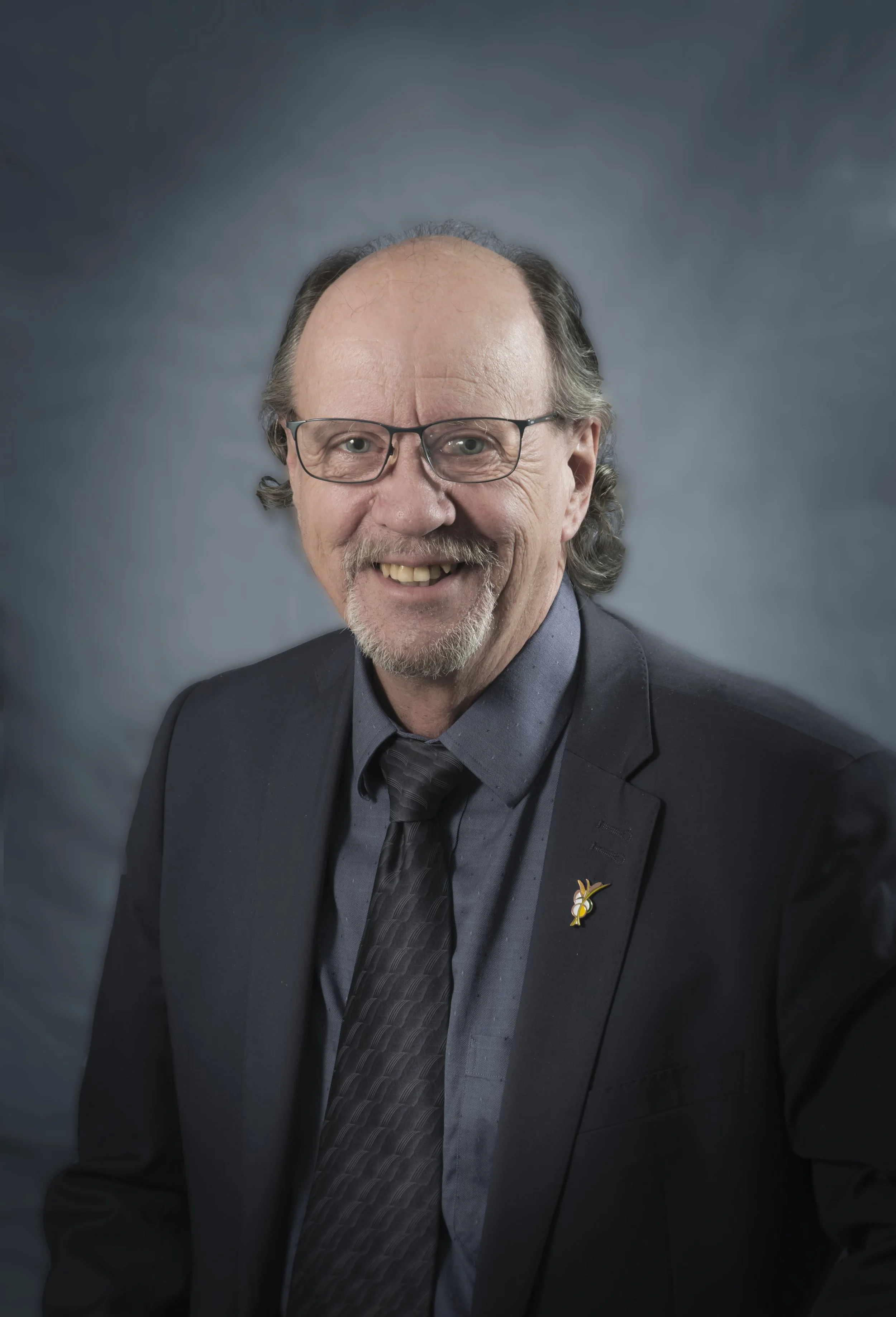 Photo d'un homme âgé portant des lunettes, avec un costume sombre, une chemise grise et une cravate, sourire, fond gris
