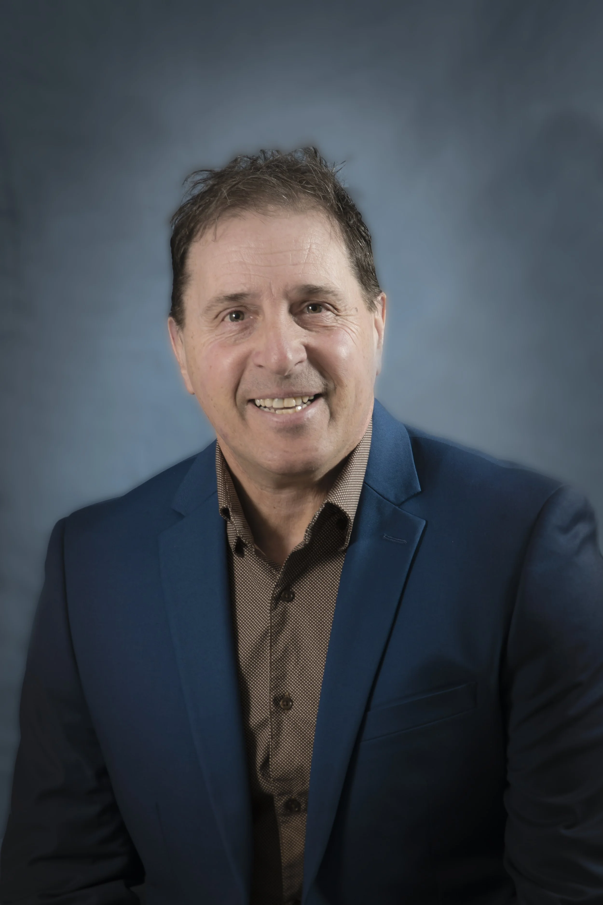 Un homme souriant en costume bleu et chemise marron clair, pose pour une photo devant un fond bleu.