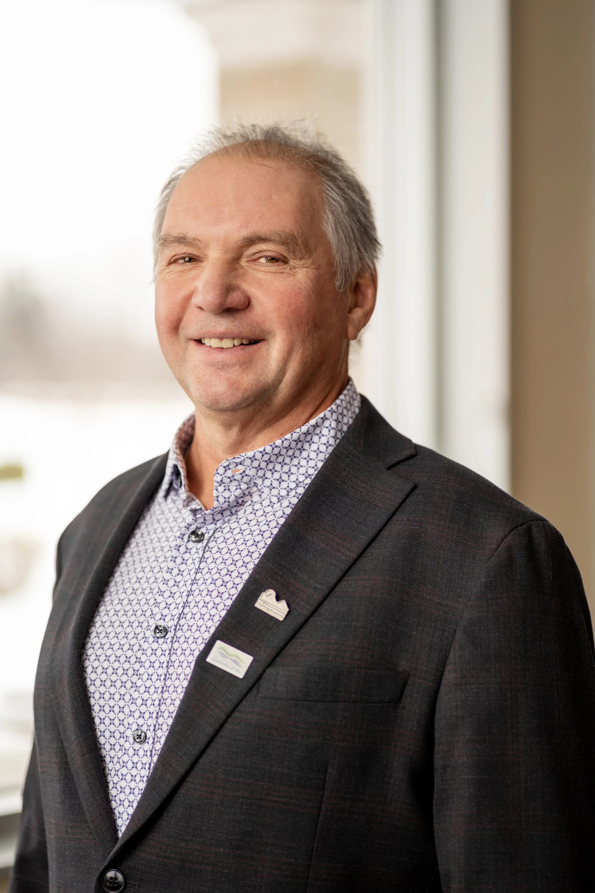 Homme d'affaires souriant portant un costume noir et une chemise à motifs, debout dans une pièce lumineuse