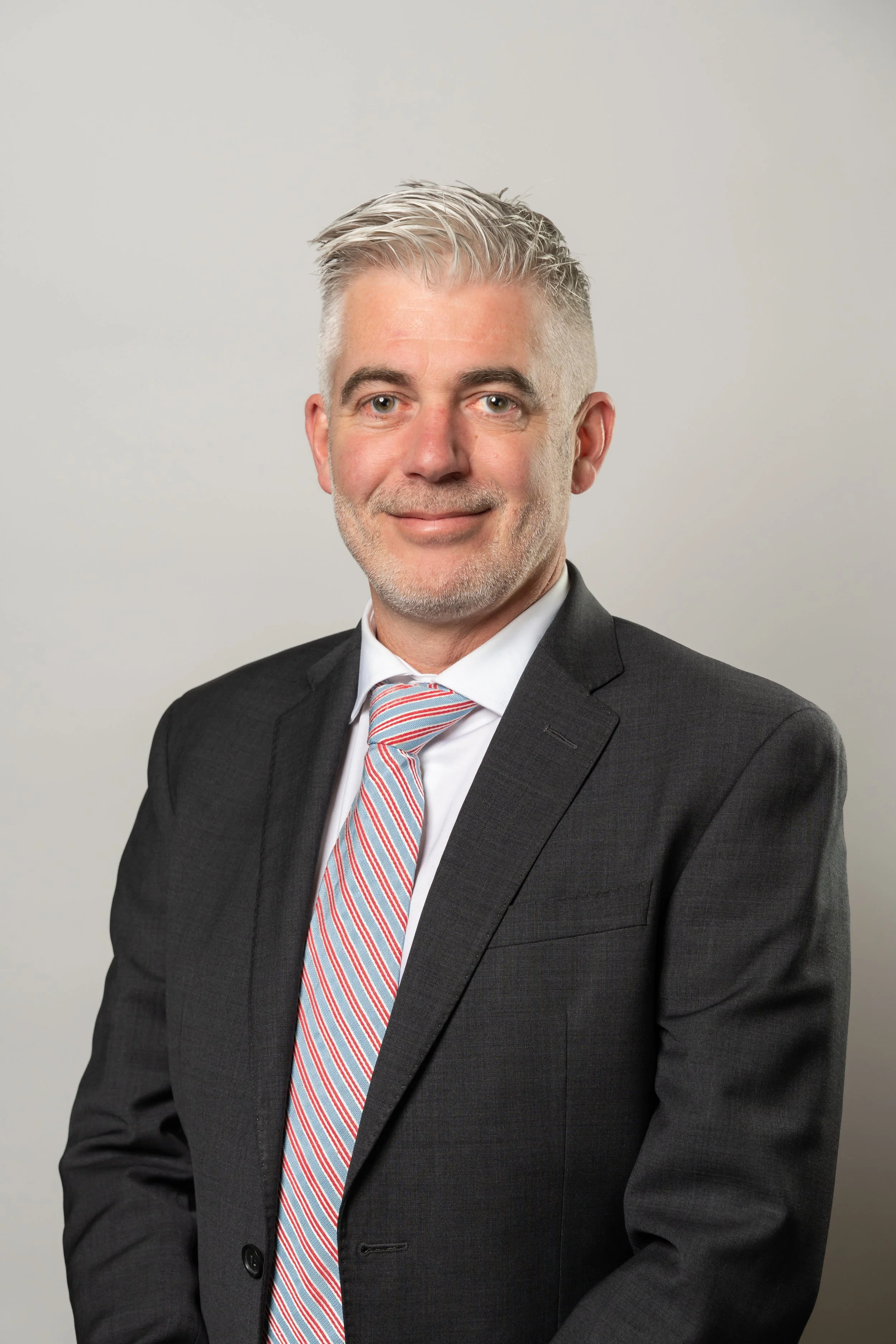 Un homme en costume noir avec une cravate à rayures rouges et blanches, souriant, contre un fond gris clair.