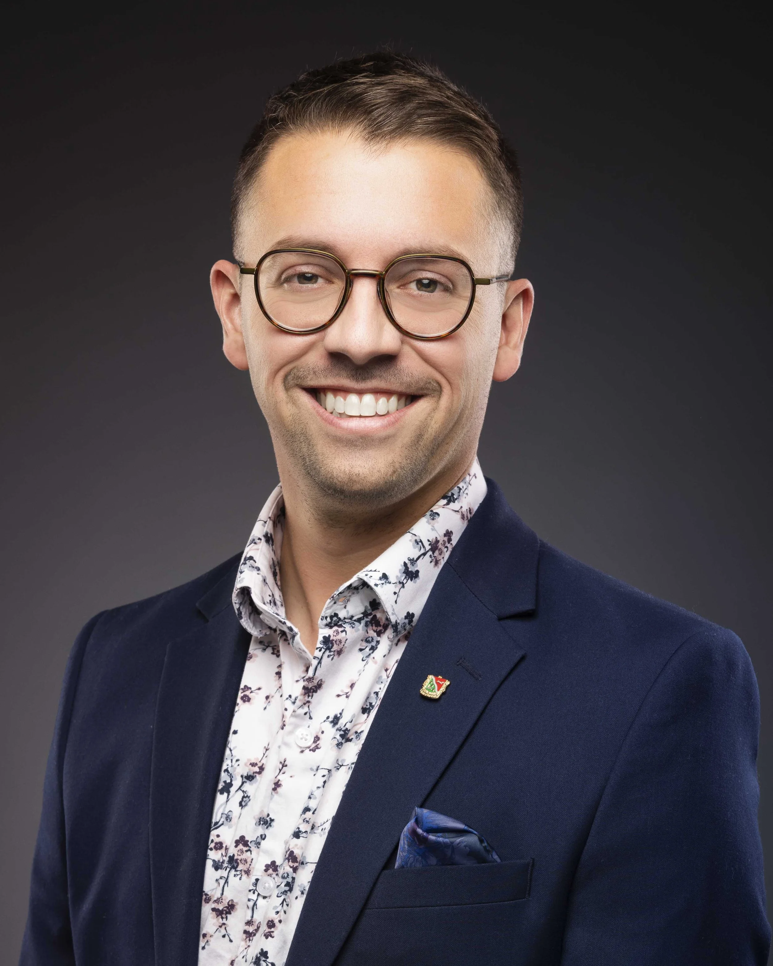 Portrait d'un homme souriant portant des lunettes, un costume bleu, une chemise à motifs floraux, et une broche sur la veste.