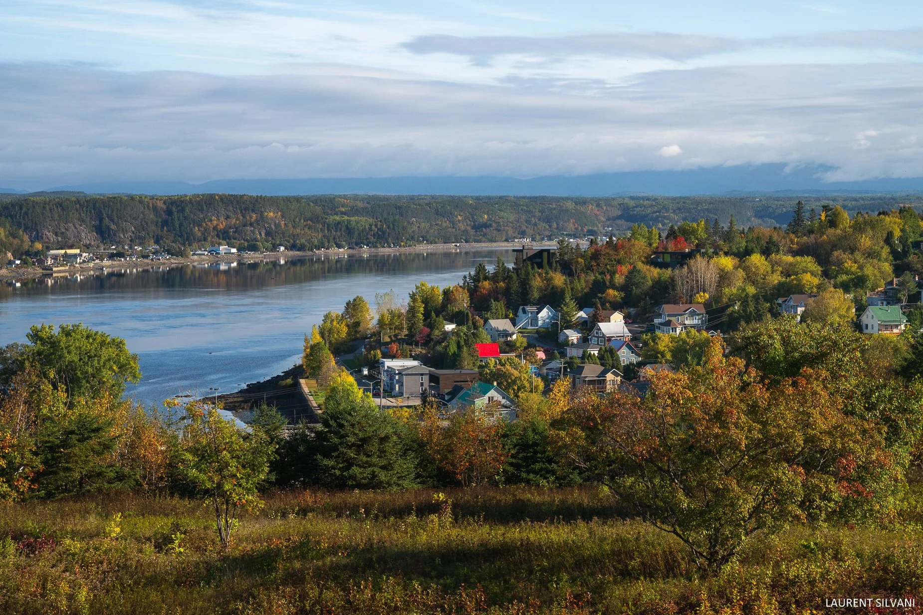 Paysage avec une rivière bordée de maisons, arbres aux couleurs automnales, montagnes au loin et ciel nuageux.