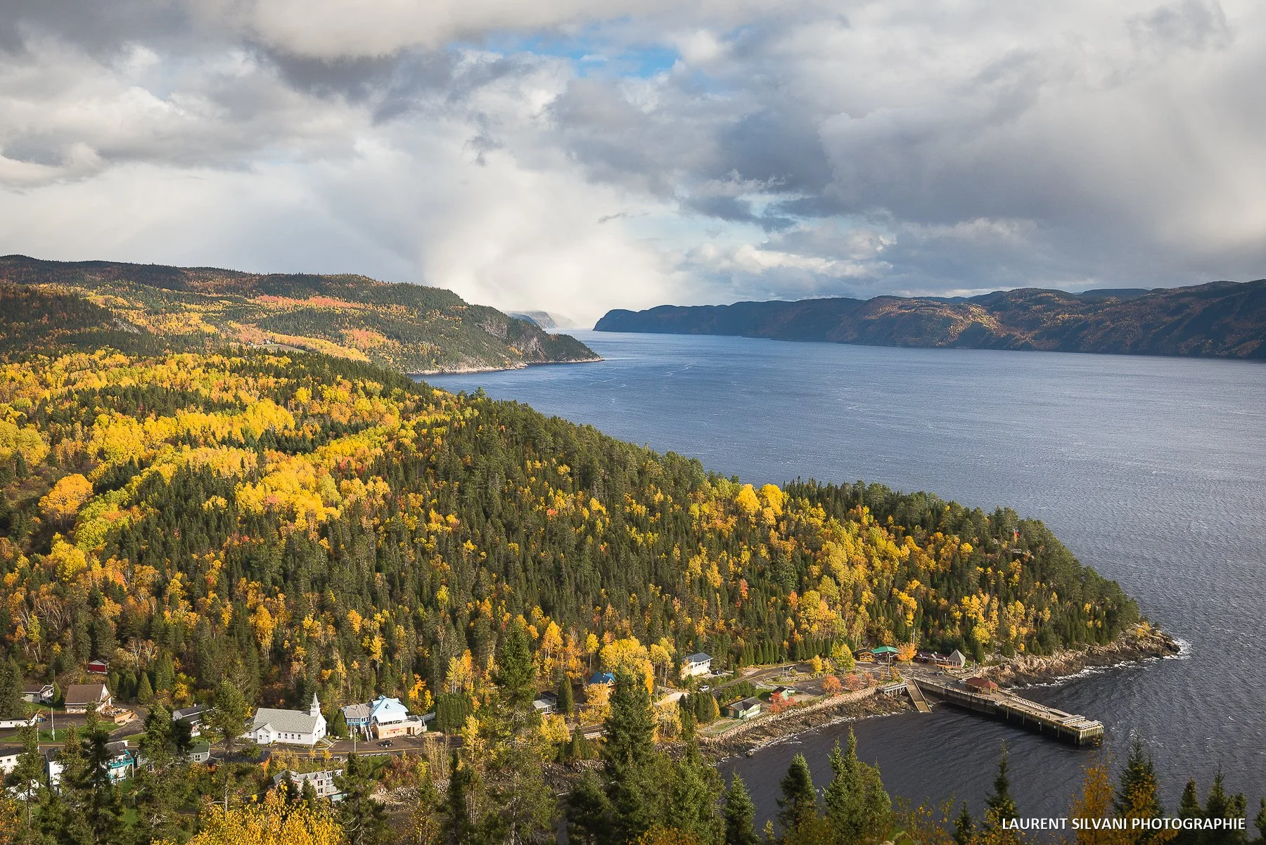 Paysage naturel avec un fleuve bordé de forêts colorées en automne, petites maisons et une jetée au bord de l'eau, ciel nuageux.