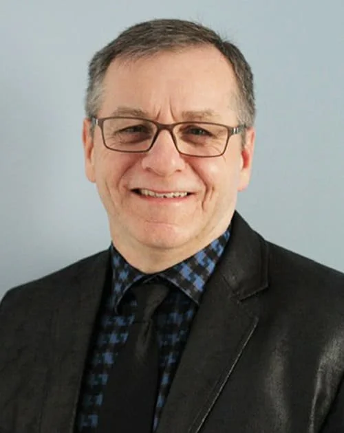 Homme souriant portant des lunettes, une chemise à carreaux bleus, une cravate et une veste noire, contre un fond neutre.