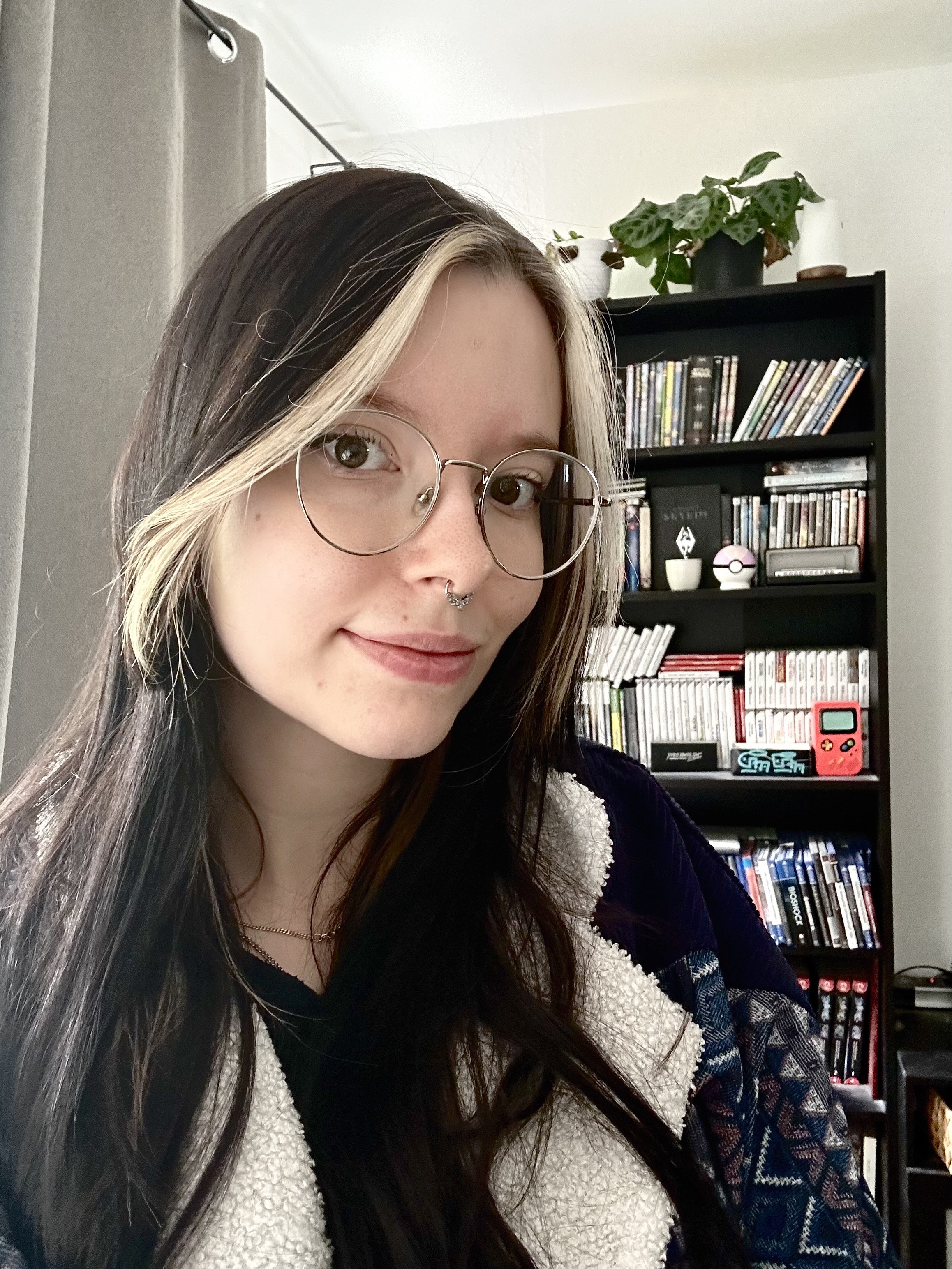 Porträt einer jungen Frau mit runder Brille und Nasenpiercing vor einem Bücherregal mit Zimmerpflanze.