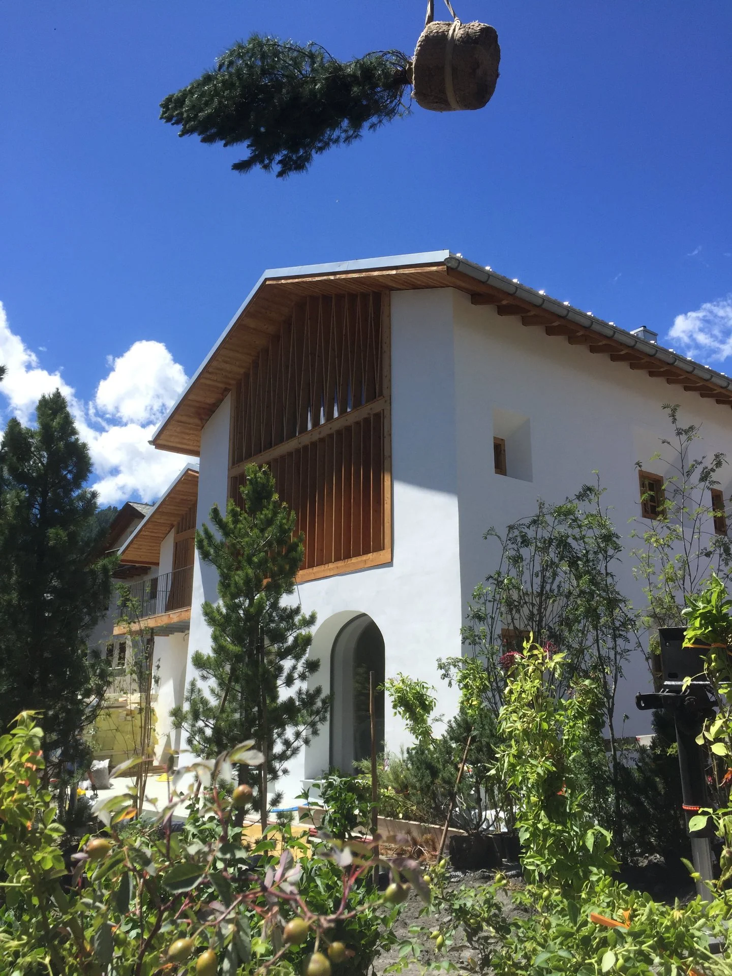 A white house with a wooden balcony and sloped roof, surrounded by green trees and plants, with a bright blue sky and some clouds in the background.