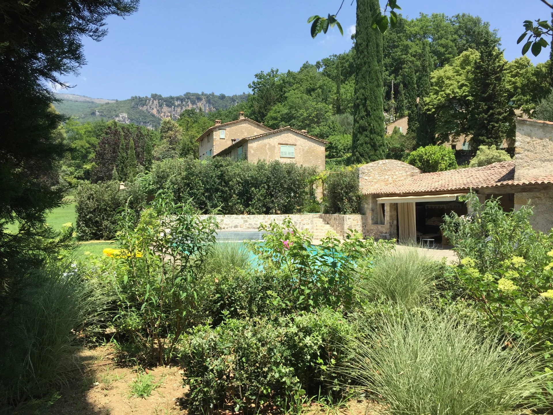 A lush garden scene with a swimming pool, stone buildings with tiled roofs, surrounded by various green trees and shrubs, set against a hillside with mountainous terrain in the background.