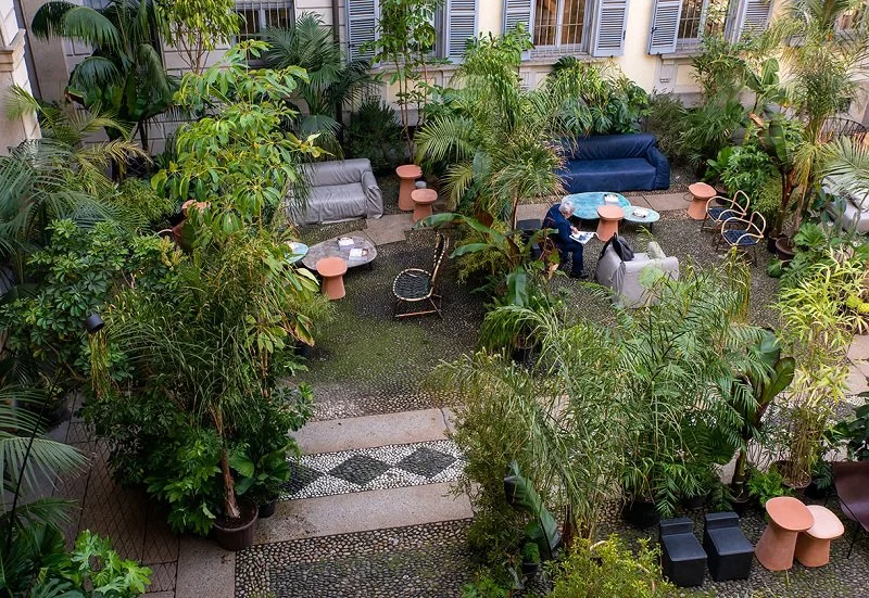 Outdoor courtyard with lush green plants and trees, seating areas with sofas and chairs, and small side tables, with two people sitting at a table.