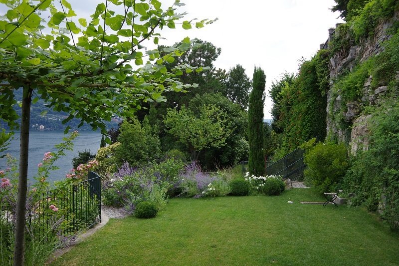 Lush garden with various trees, shrubs, and flowering plants next to a river, with a grassy lawn, stone wall, and a bench.