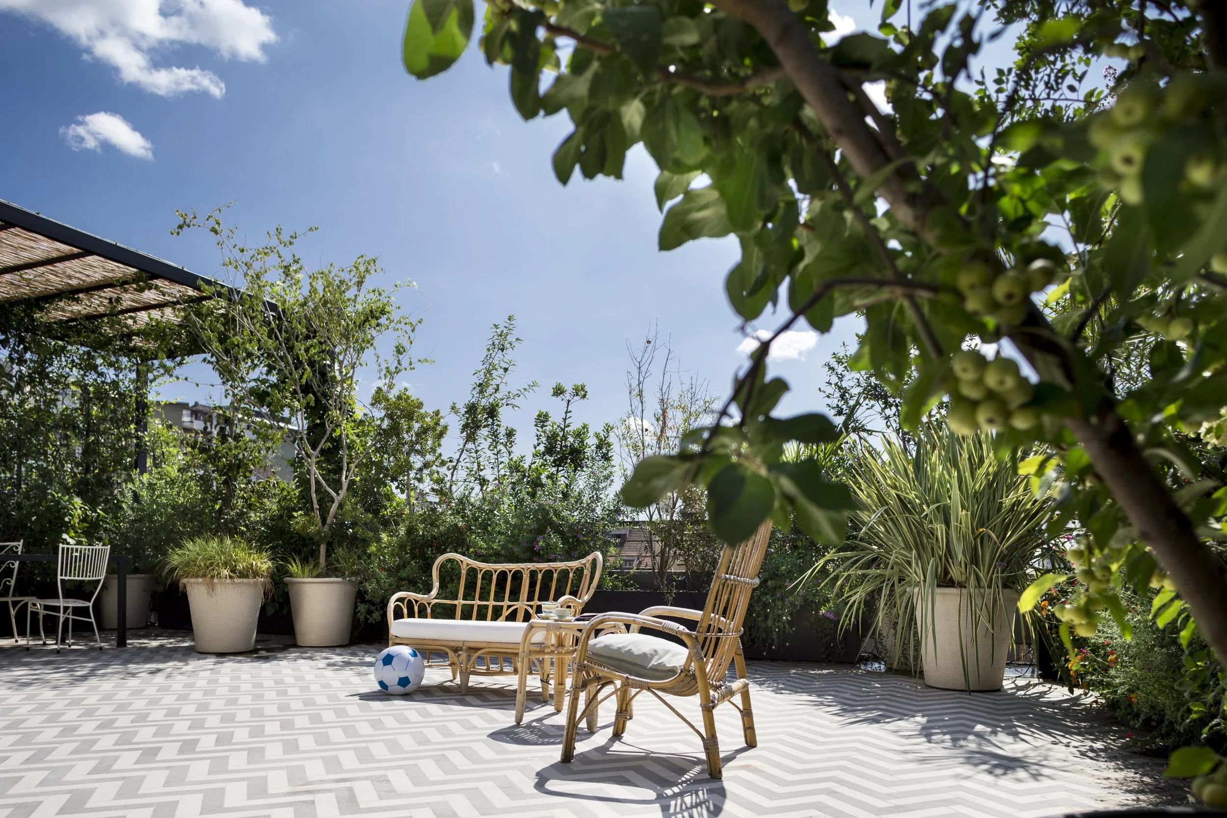 Outdoor patio with wicker furniture, potted plants, a soccer ball, and lush greenery under a bright blue sky.