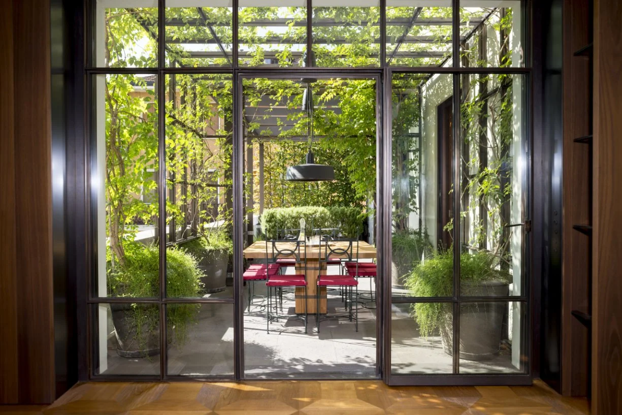 View through glass doors into a sunlit outdoor patio with a wooden dining table, six chairs with red cushions, surrounded by potted plants and greenery, with a pergola and trees overhead.