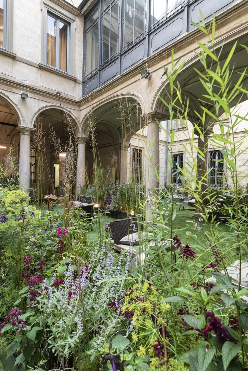 An indoor garden courtyard with lush plants and flowers, surrounded by arched hallways and windows of a classical building.