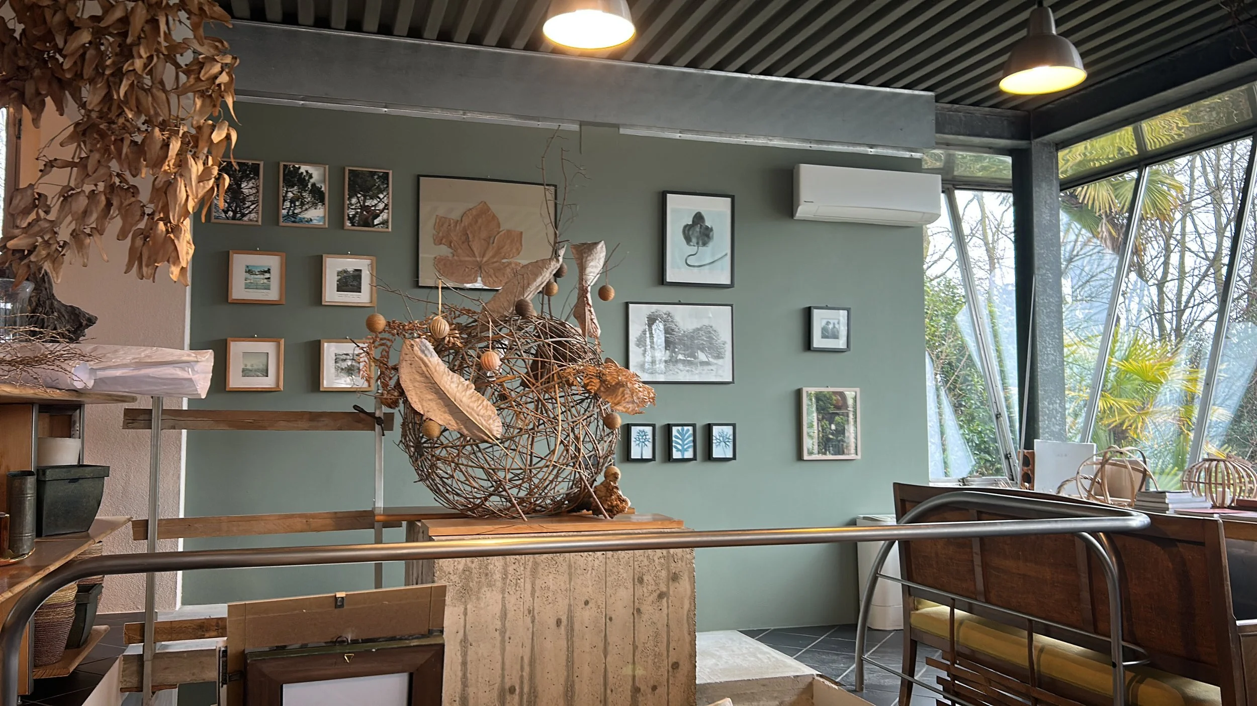 Interior of a modern room with a green wall decorated with multiple framed pictures, a large window showing trees outside, and a table with decorative dried branches and leaves.