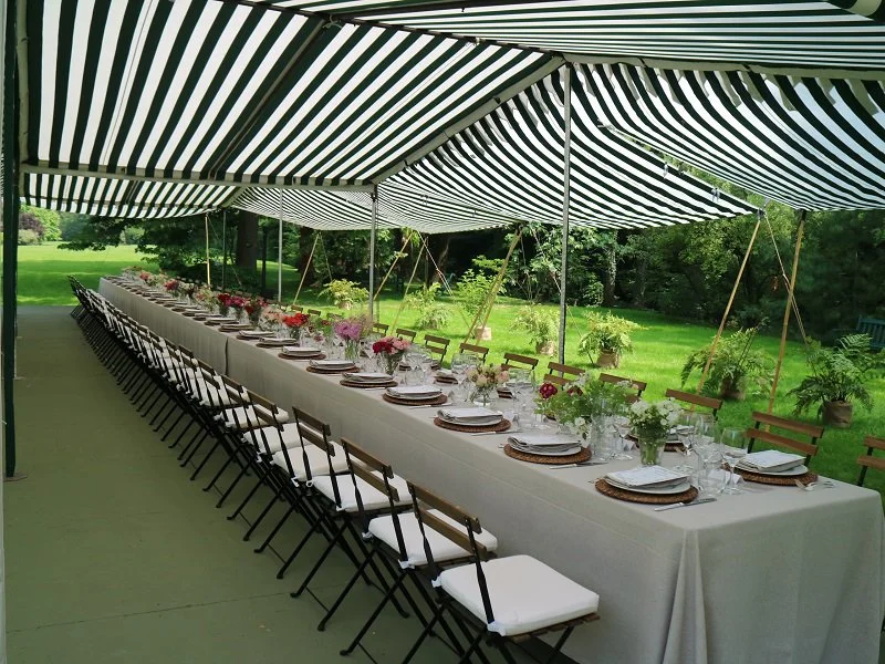 Long outdoor banquet table set under a striped canopy with floral centerpieces, plates, glasses, and utensils, surrounded by chairs on a lush green lawn.