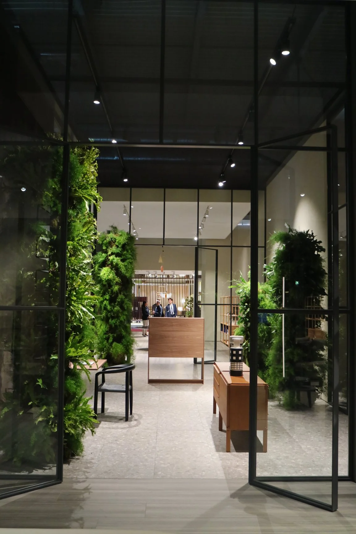 View of a modern office lobby with glass doors, green plants, a small wooden table, and people in the background.