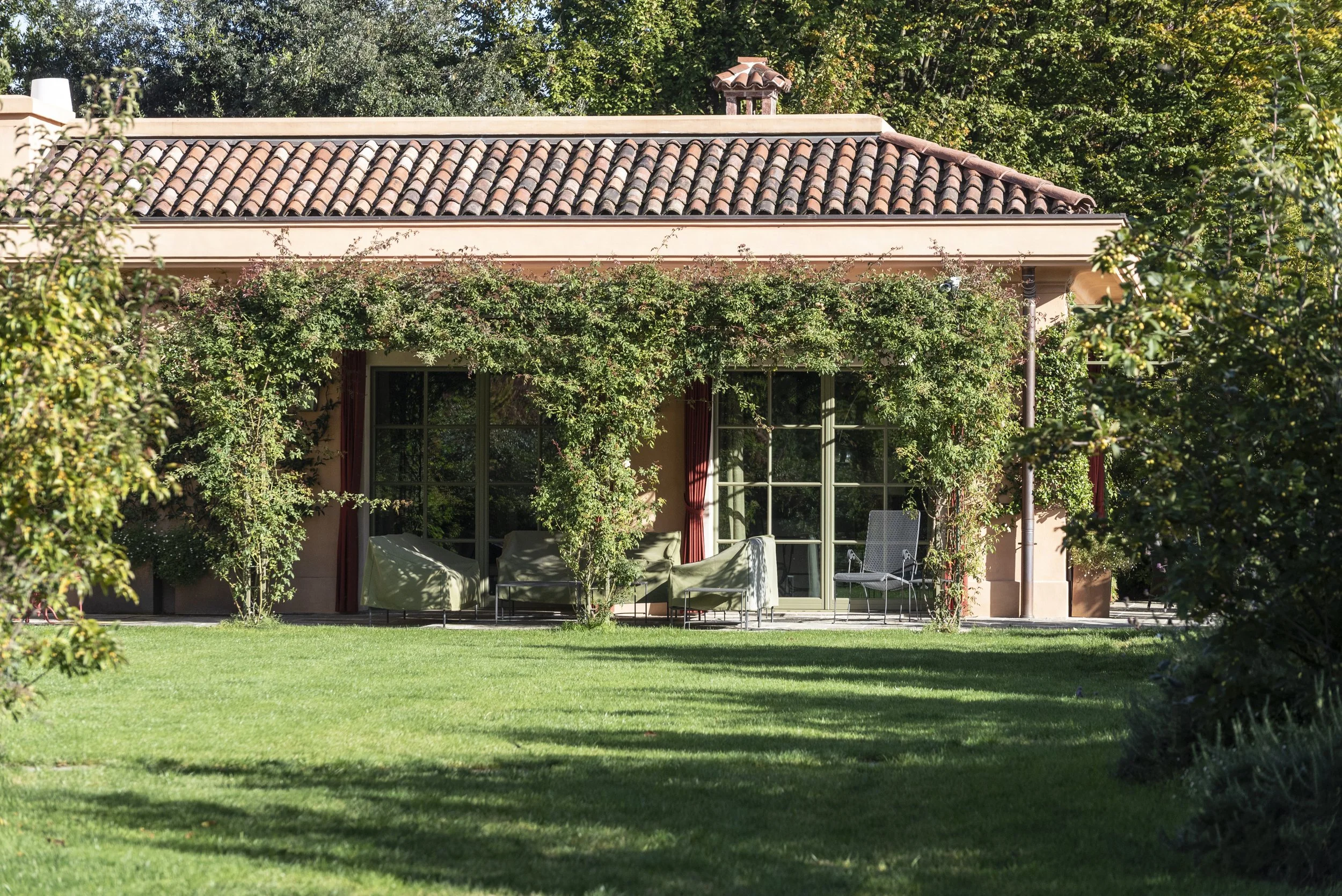 Front view of a house with large glass doors, surrounded by green vines and trees, with a well-maintained lawn in the foreground.