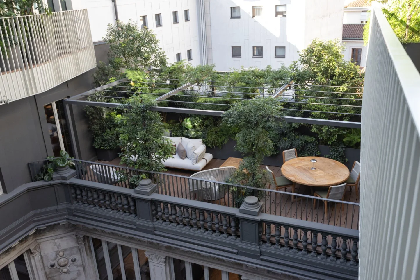 A small outdoor patio with green plants, a white sofa with pillows, a round wooden table with four chairs, and a balcony railing, surrounded by a modern building facade.