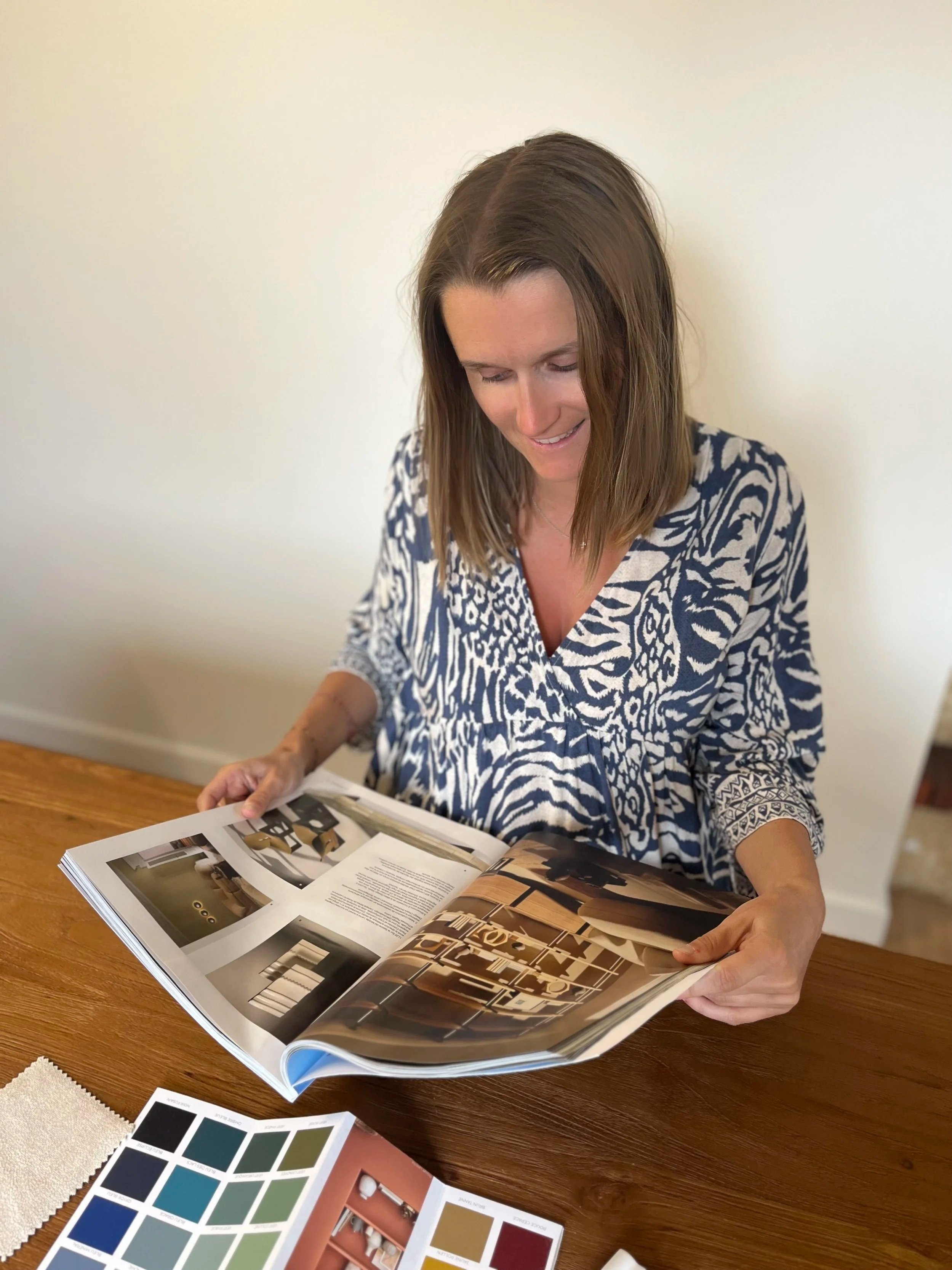 Femme regardant un magazine d'intérieur ou de design, posée sur une table en bois, avec des échantillons de tissu ou de peinture à proximité.