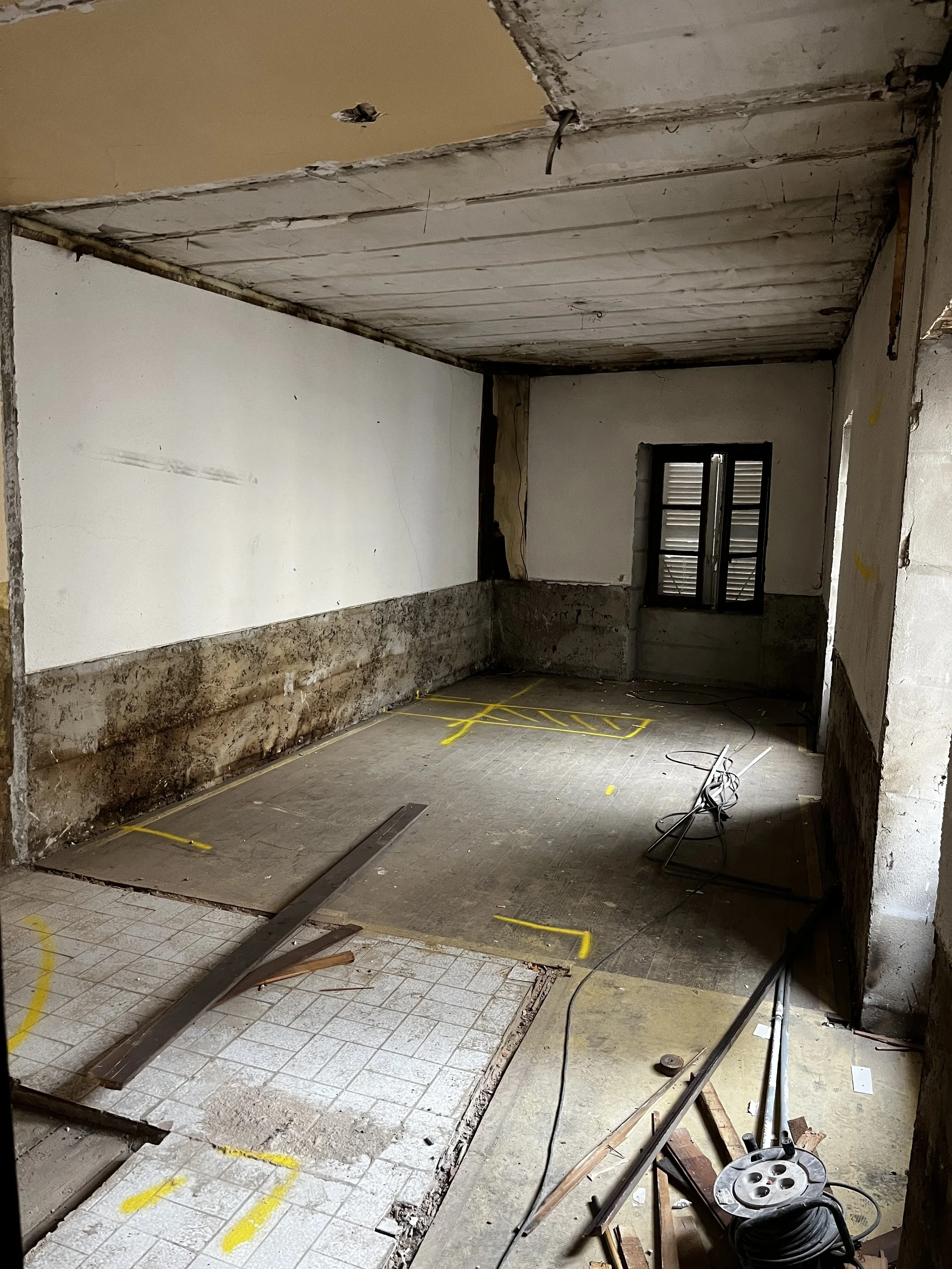 Pièce en cours de rénovation avec sol partiellement démoli, murs en cours de reconstruction, fenêtres à l'arrière, outils et câbles électriques.»
