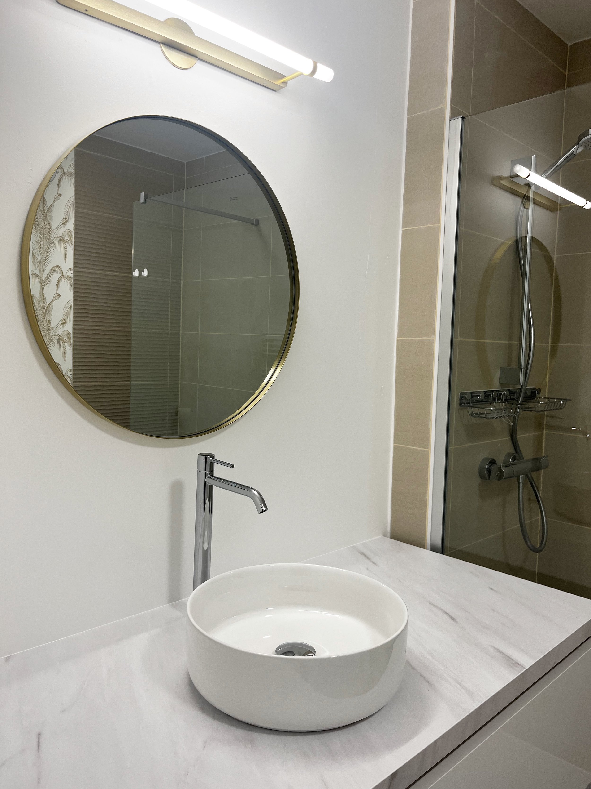 Une salle de bain moderne avec un lavabo rond blanc sur un comptoir en marbre, un miroir rond au-dessus, une lumière horizontale au-dessus du miroir, et une douche avec porte en verre et paroi en beige à l'arrière.