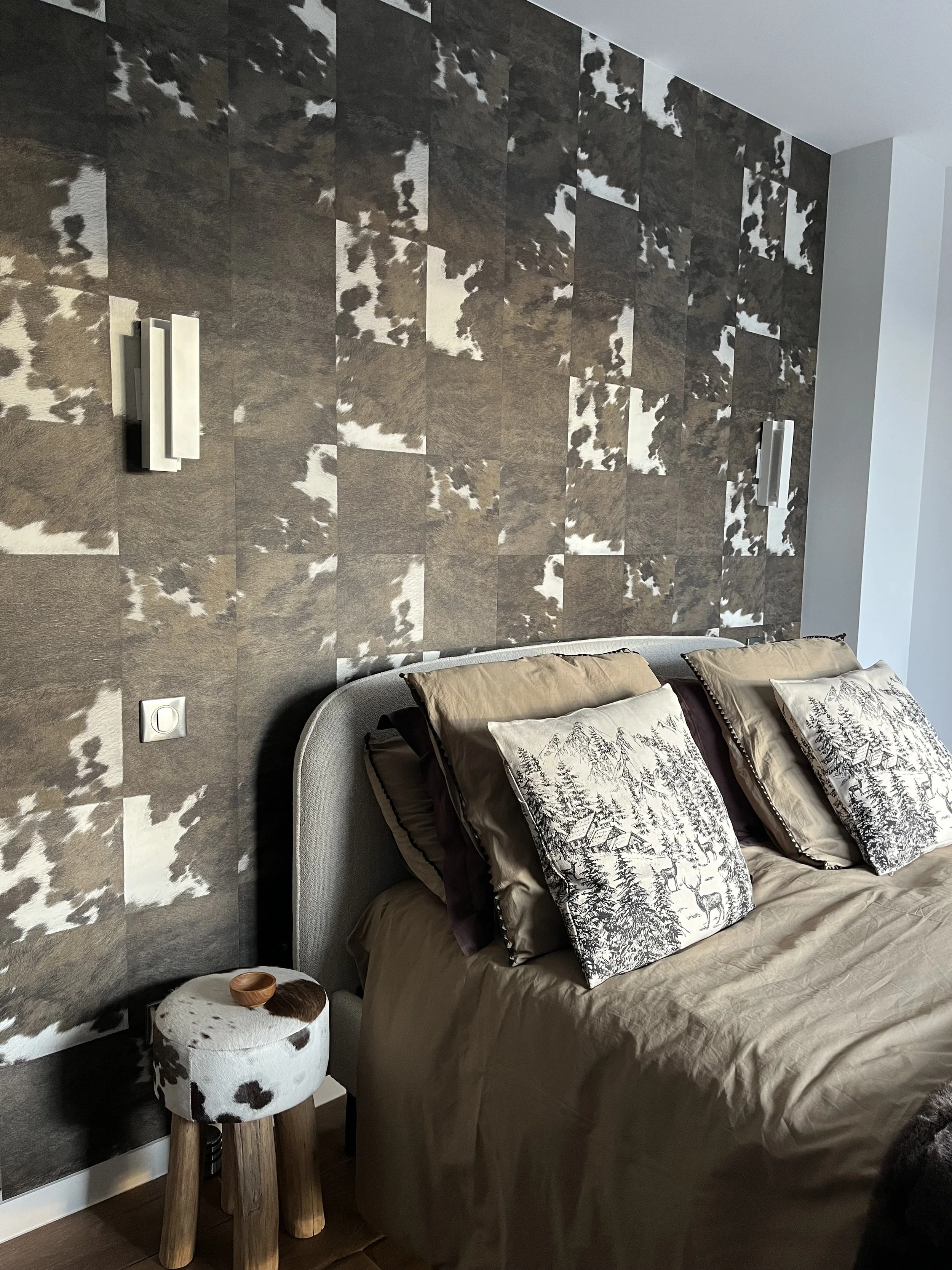 Chambre à coucher avec un mur en tissu ou en papier à motif cowhide, un lit avec une tête de lit gris, des oreillers avec des motifs de forêt, une petite table en bois avec un coussin en peau de vache et un bol en bois.
