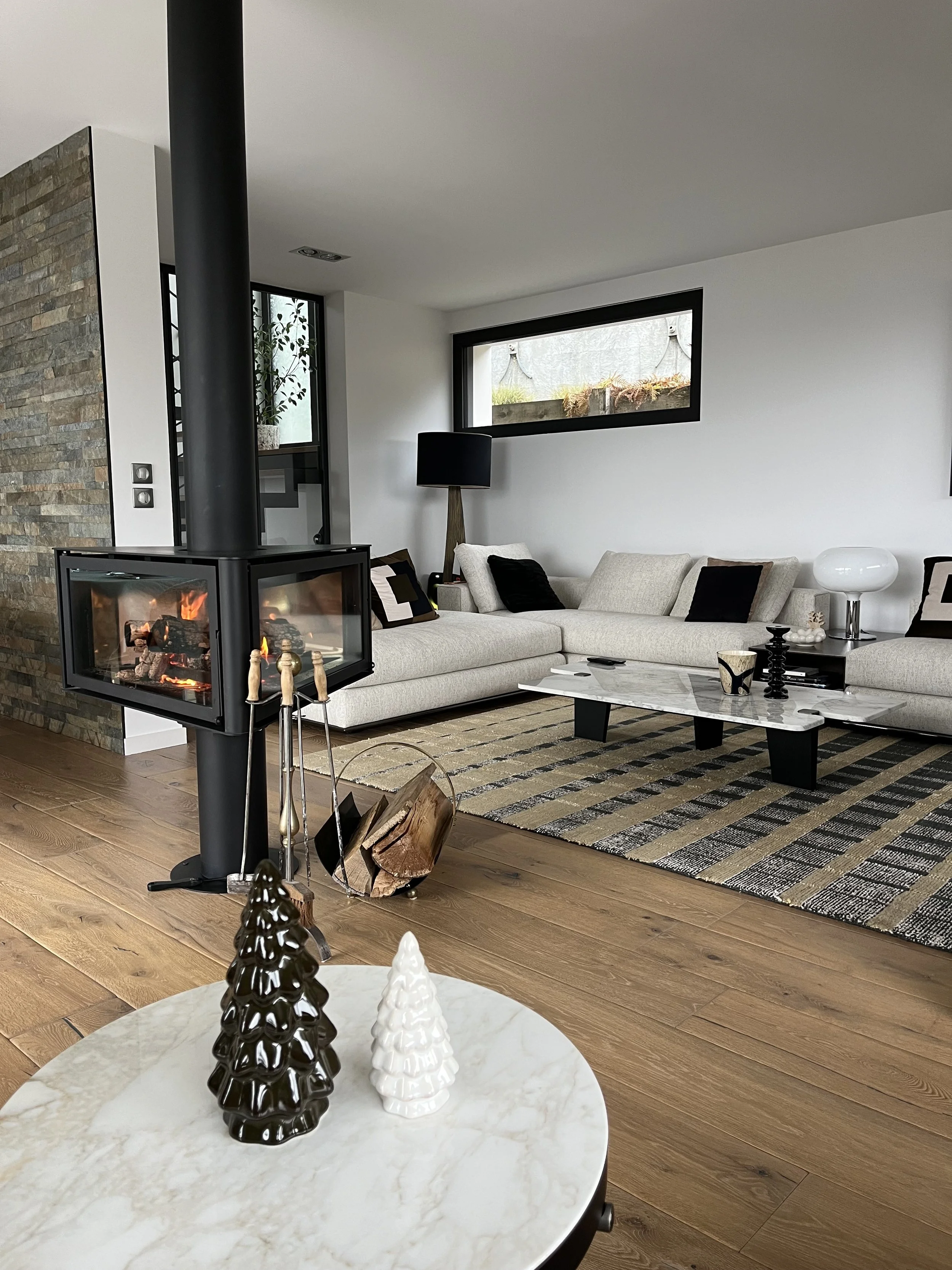 Un salon moderne avec canapé beige, table basse en marbre, cheminée moderne en verre, planche de bois au sol, et décorations de Noël en céramique en forme de sapin sur une table