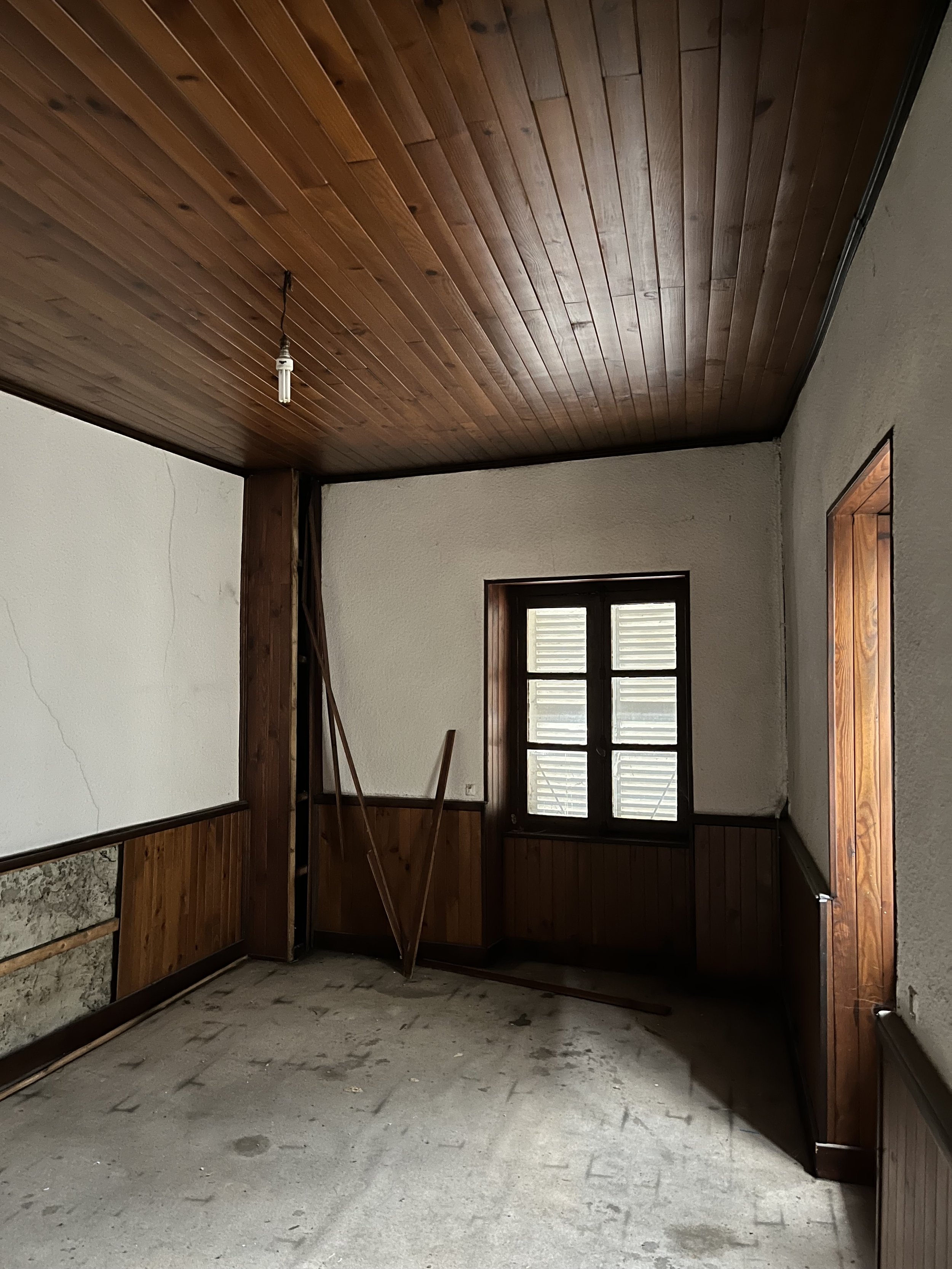 Pièce vide avec plafond en bois, murs en plâtre, fenêtre à gauche, et sol sans revêtement, en cours de rénovation.
